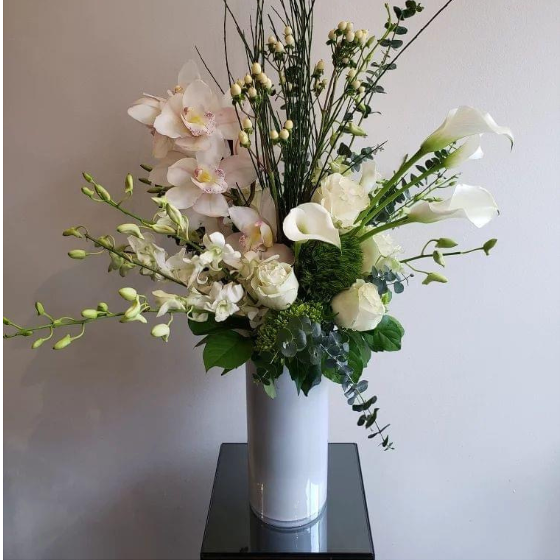Elegant floral arrangement of white orchids, roses, and calla lilies in a tall white vase with green foliage