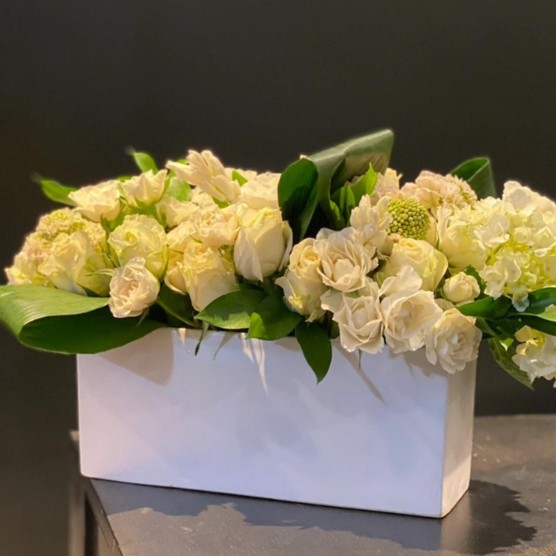 Symmetrical floral arrangement of cream roses and hydrangeas in a rectangular white vase with green leaves on dark background