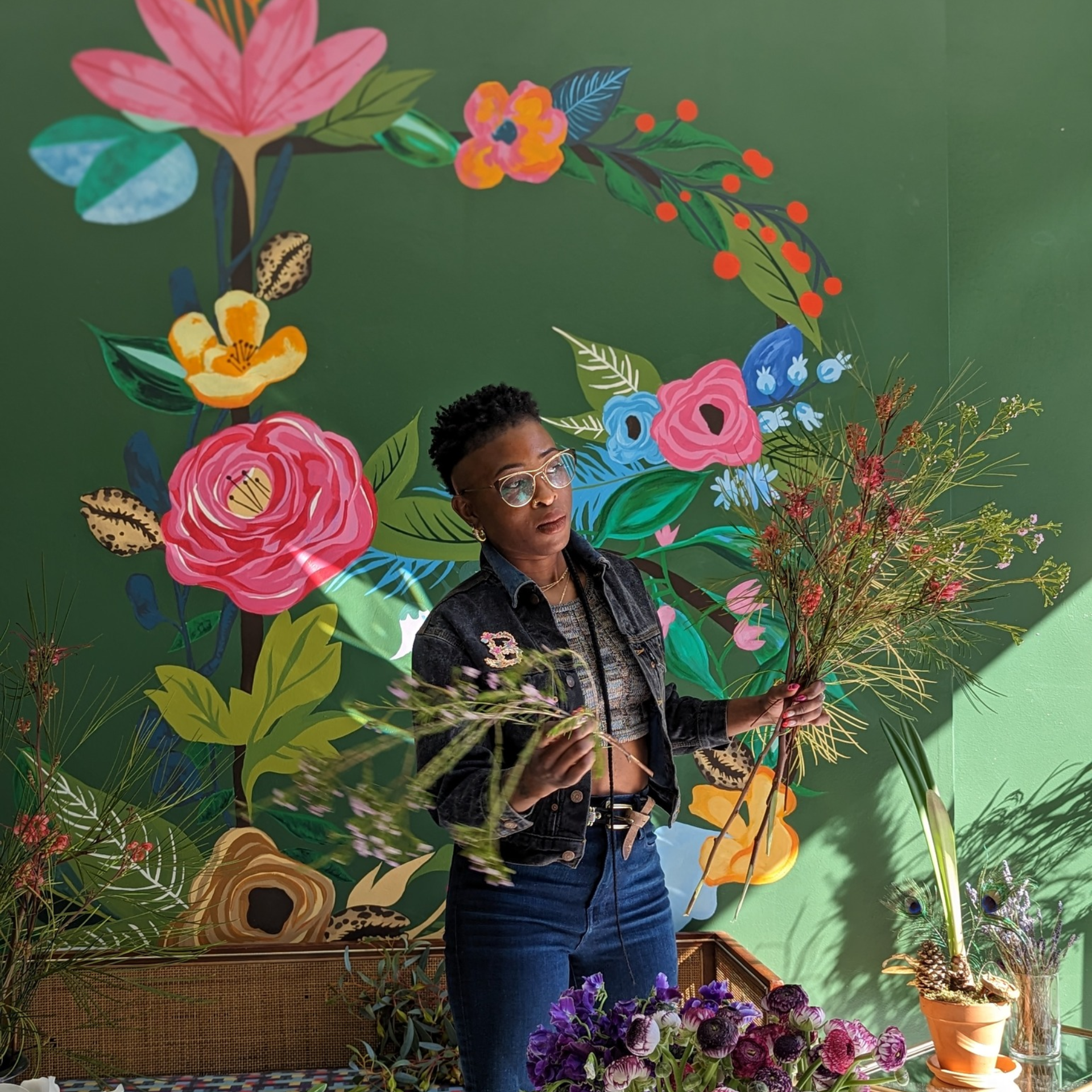 Person holding plants in front of colorful floral mural with potted flowers on table