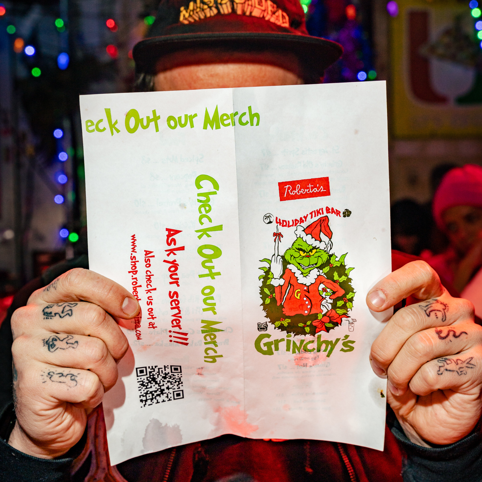 Person holding a festive flyer for Roberta’s Holiday Tiki Bar Grinchy’s, with colorful lights behind