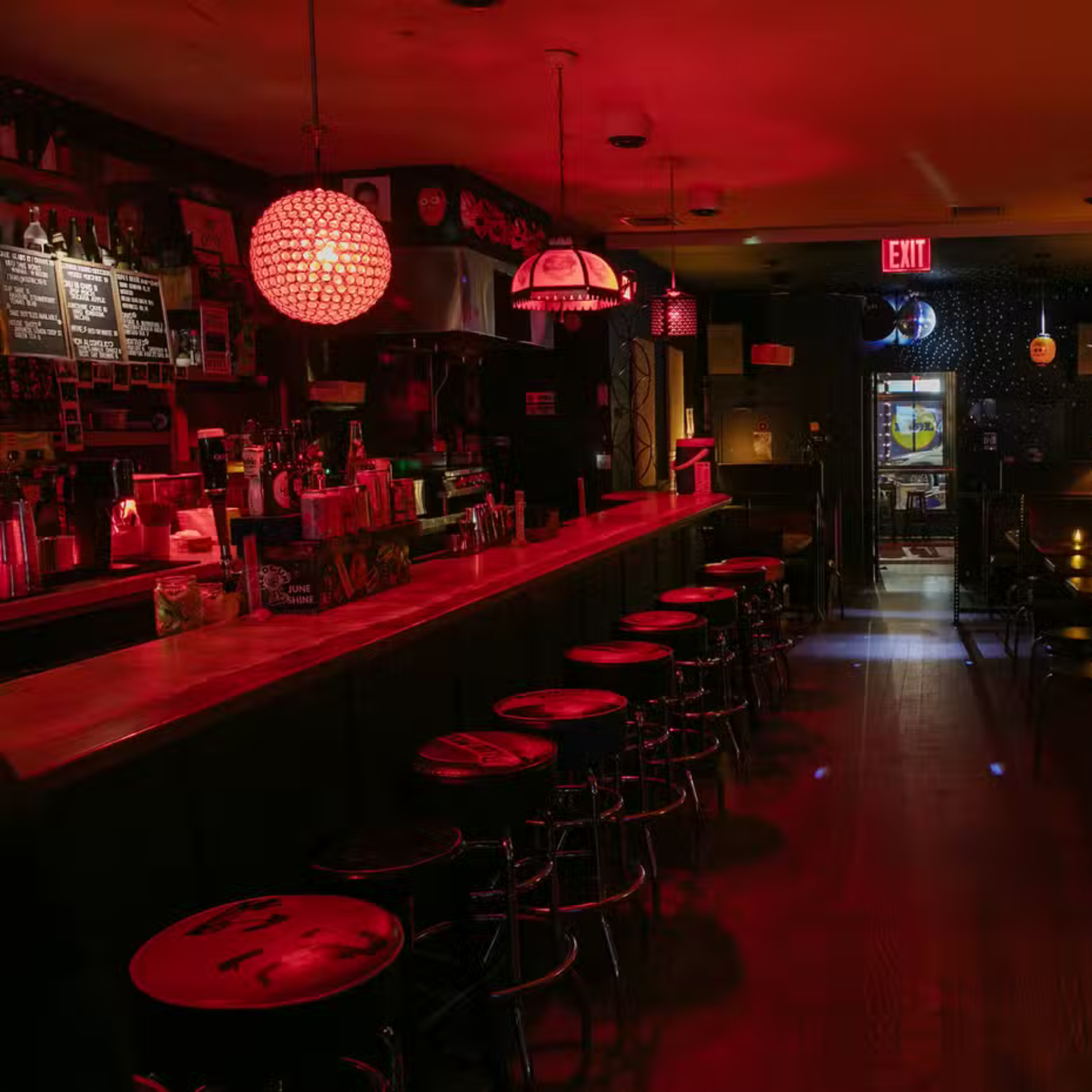 Dimly lit bar interior with red lighting, wooden counter, and hanging pendant lights above empty stools