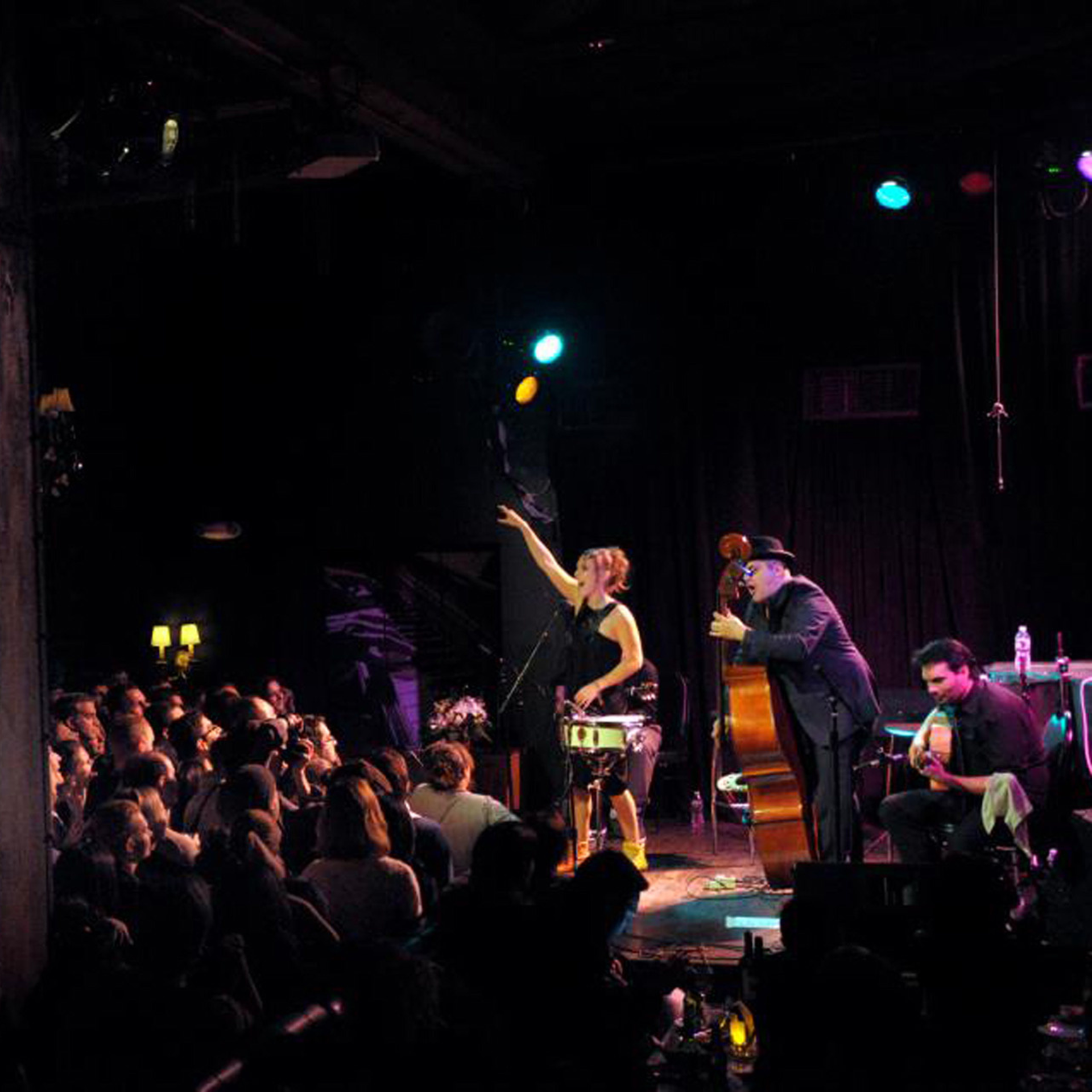 Live jazz performance on stage at a nightclub with a female vocalist, a double bassist, and two guitarists, audience seated and enjoying the show under colorful stage lighting