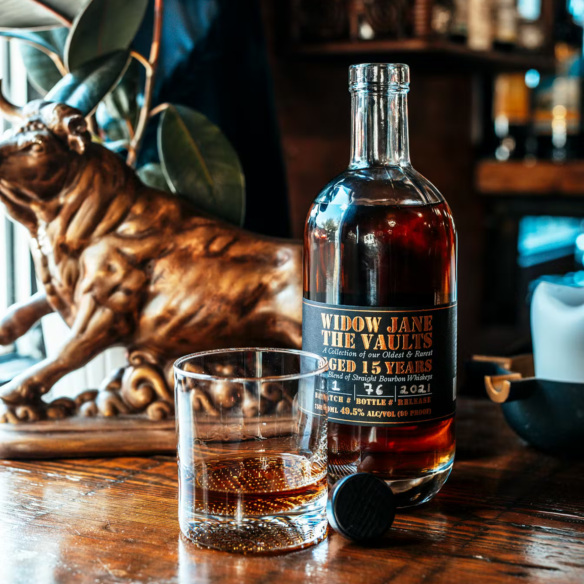 - A bottle of Widow Jane “The Vaults” bourbon and a glass of whiskey on a wooden table, with a bronze bull sculpture in the background