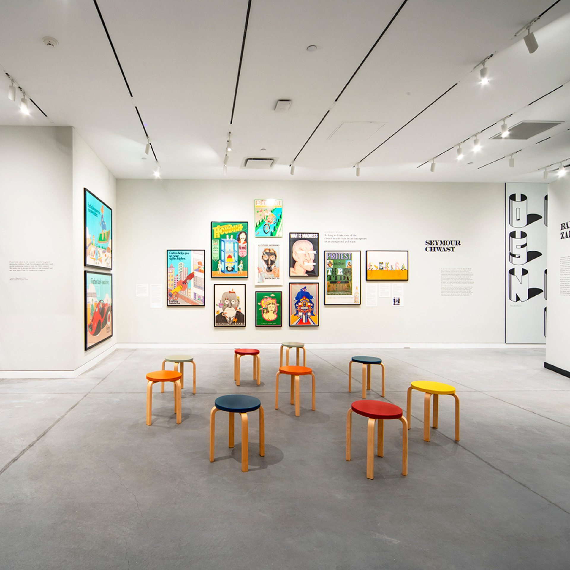 Contemporary art gallery with colorful framed artwork on white walls, modern ceiling lighting, and wooden stools with vibrant upholstered seats arranged on a polished concrete floor