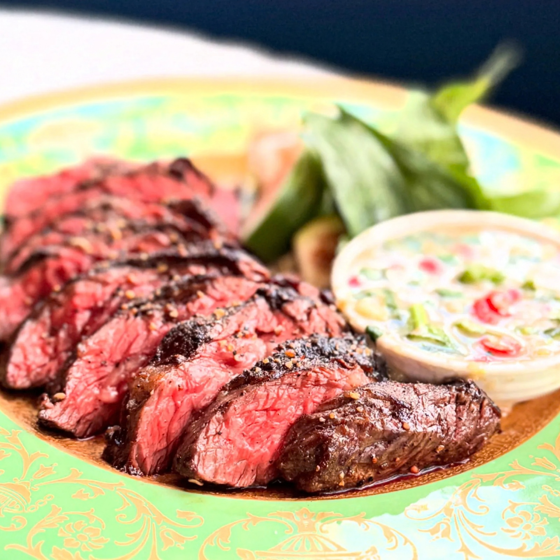 Plate of sliced medium-rare steak with dipping sauce and fresh herbs on decorative plate