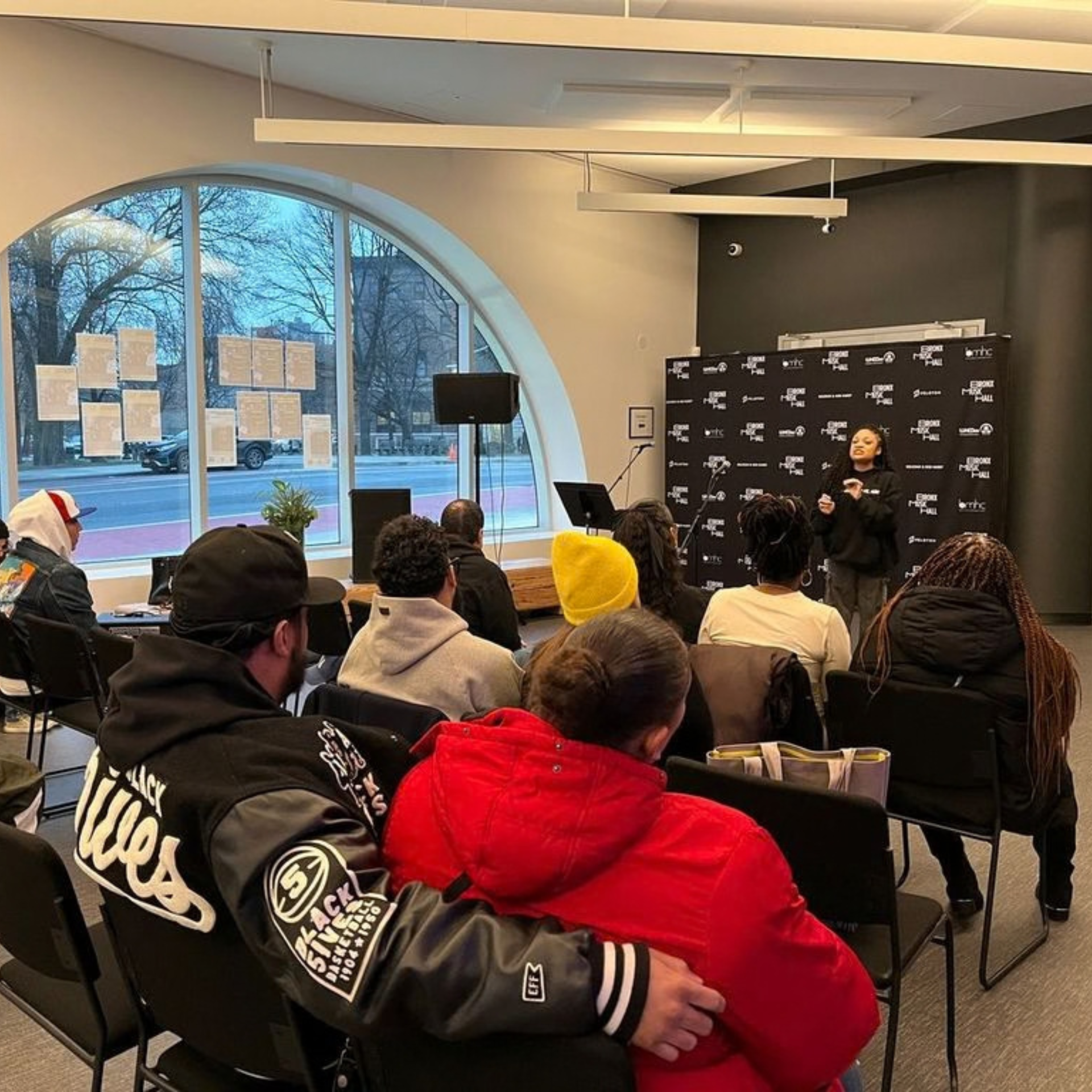 Audience listens to a speaker at an indoor event with logos on backdrop and winter scene outside window