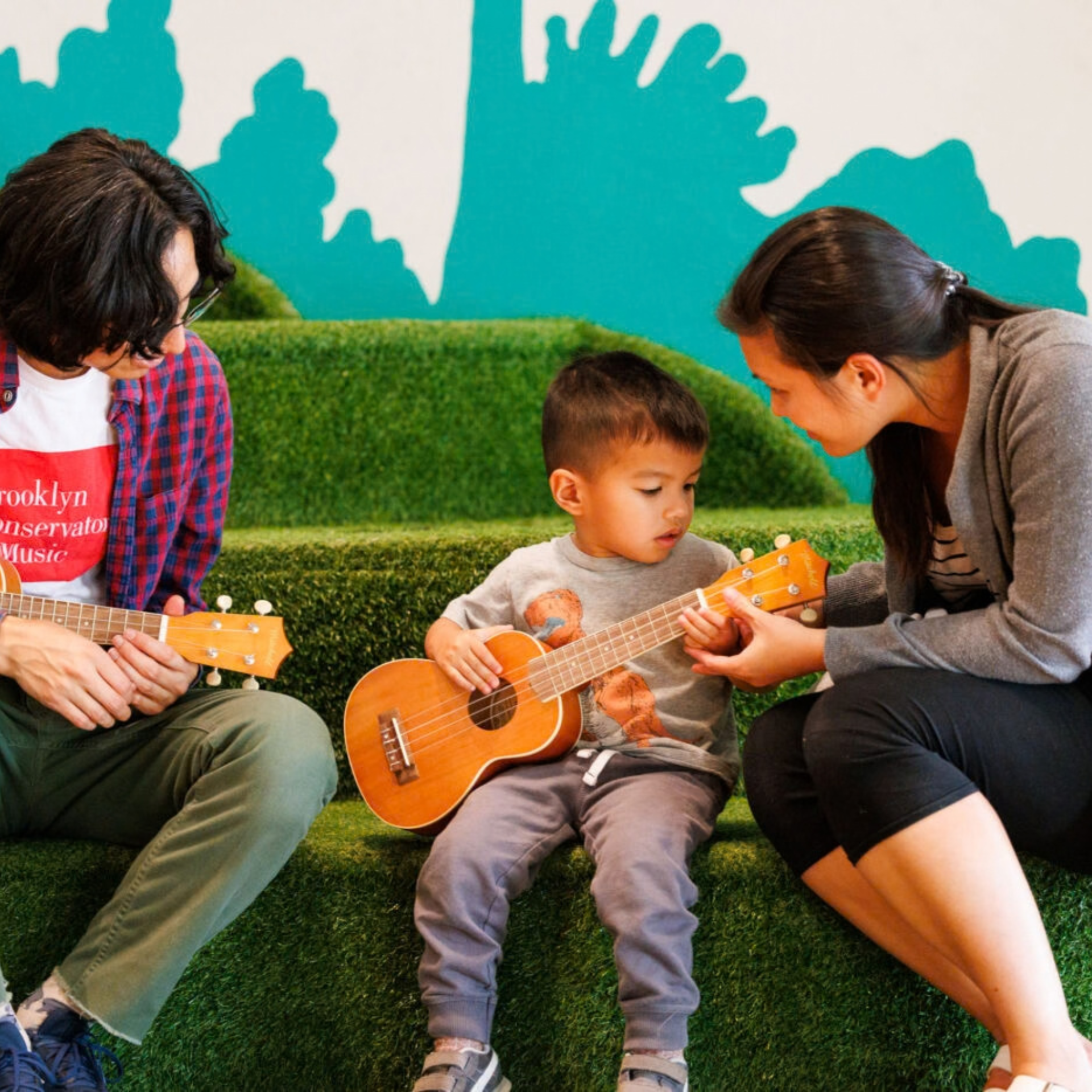 Child with ukulele sits on grass steps, guided by two adults during a music lesson