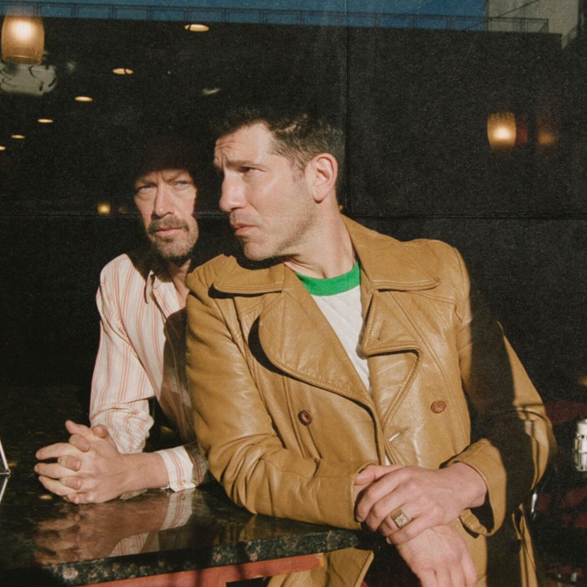 Two people sit at a café table, sunlight highlighting their faces against a dark interior