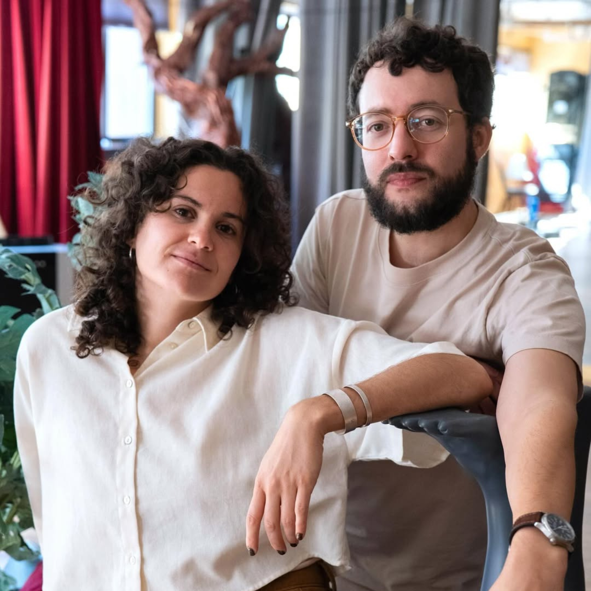Two people with curly hair standing indoors together, one in a white shirt and the other in a beige t-shirt with glasses, against a backdrop of red curtains and artistic décor