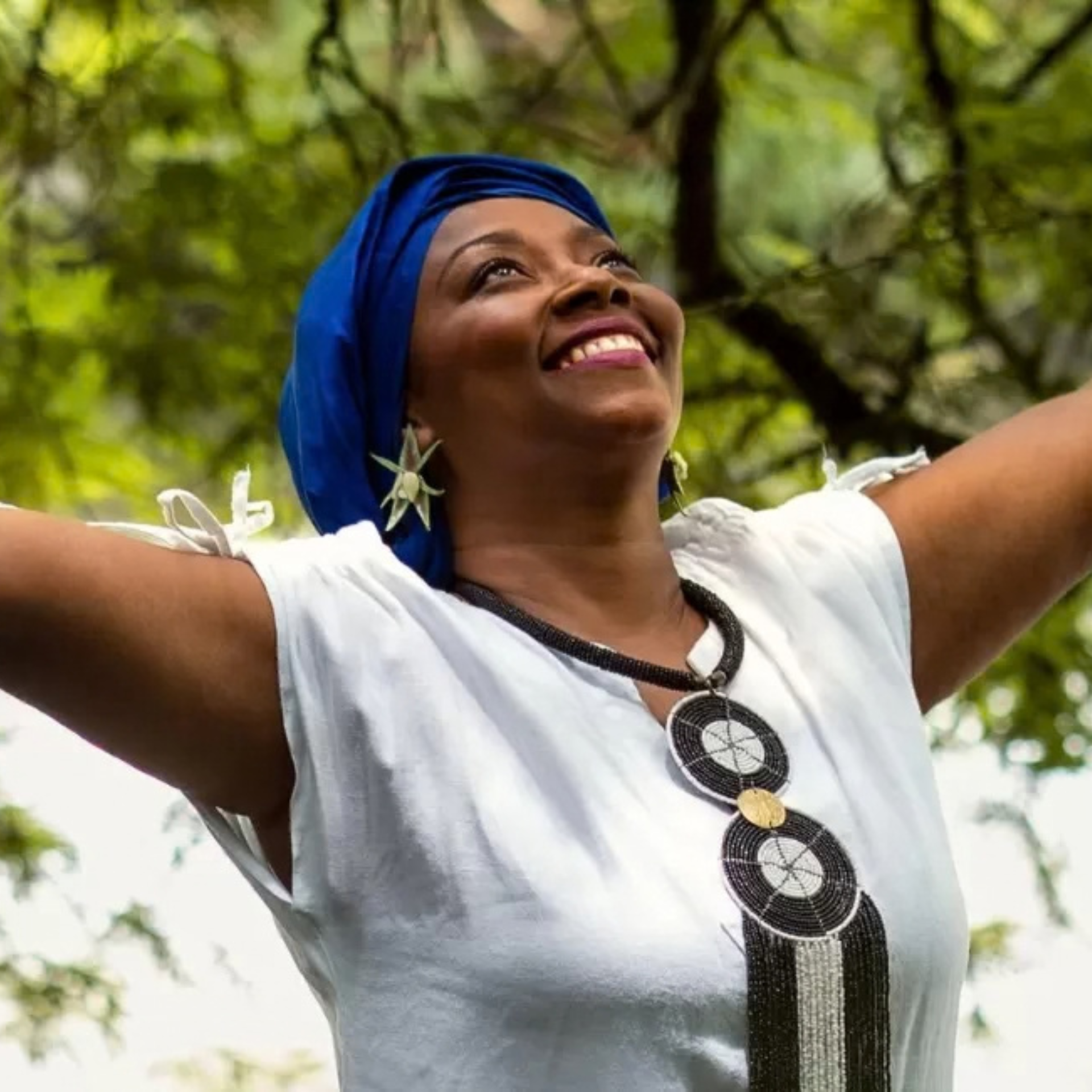 Person outdoors with arms raised, wearing blue headwrap, jewelry, and white top against green foliage