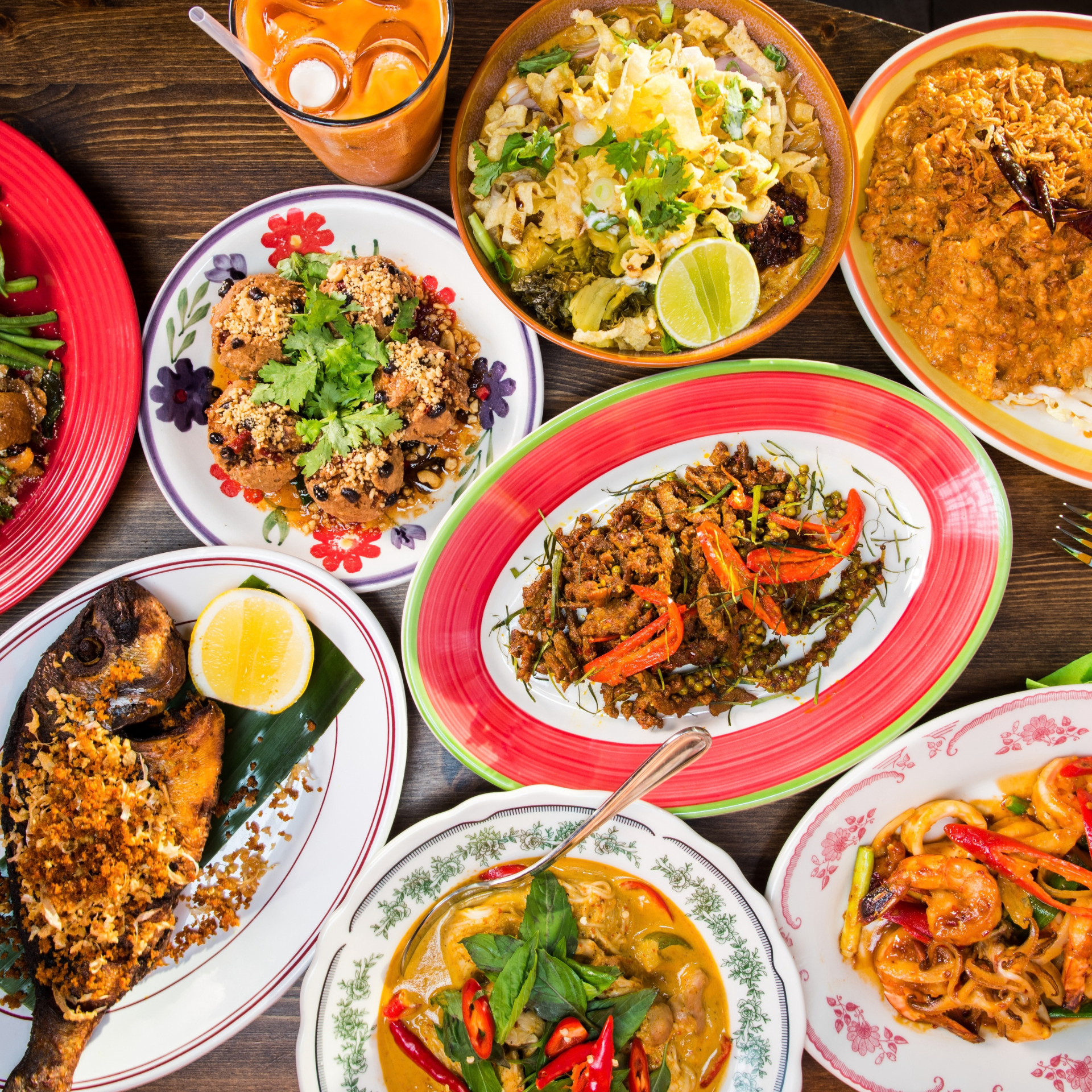 Wooden table filled with colorful Thai dishes, including fried fish, curry, stir-fry, and iced tea