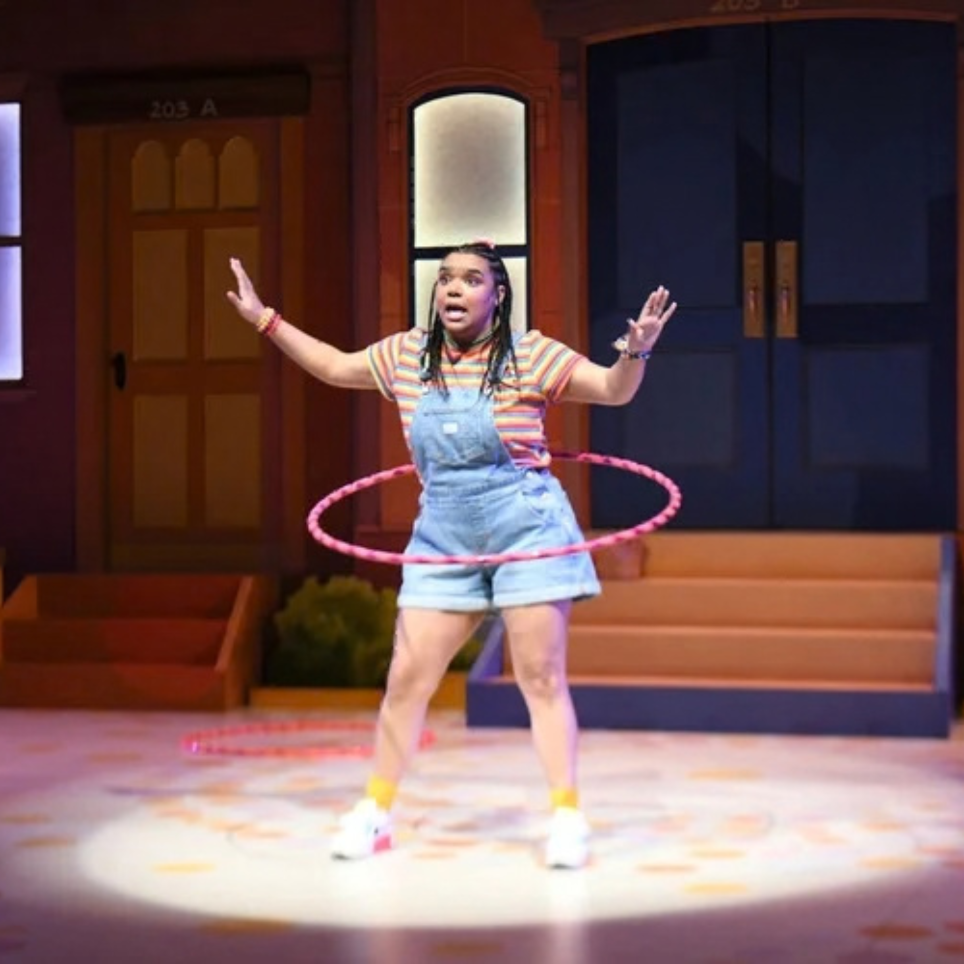 Performer on stage in striped shirt and overalls using a pink hula hoop, with doorways labeled 203 A and 203 B in background