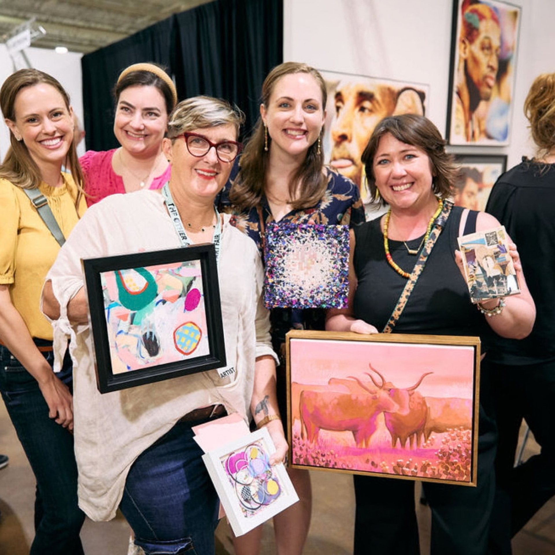 Five women smiling indoors, each holding colorful artwork at an exhibition