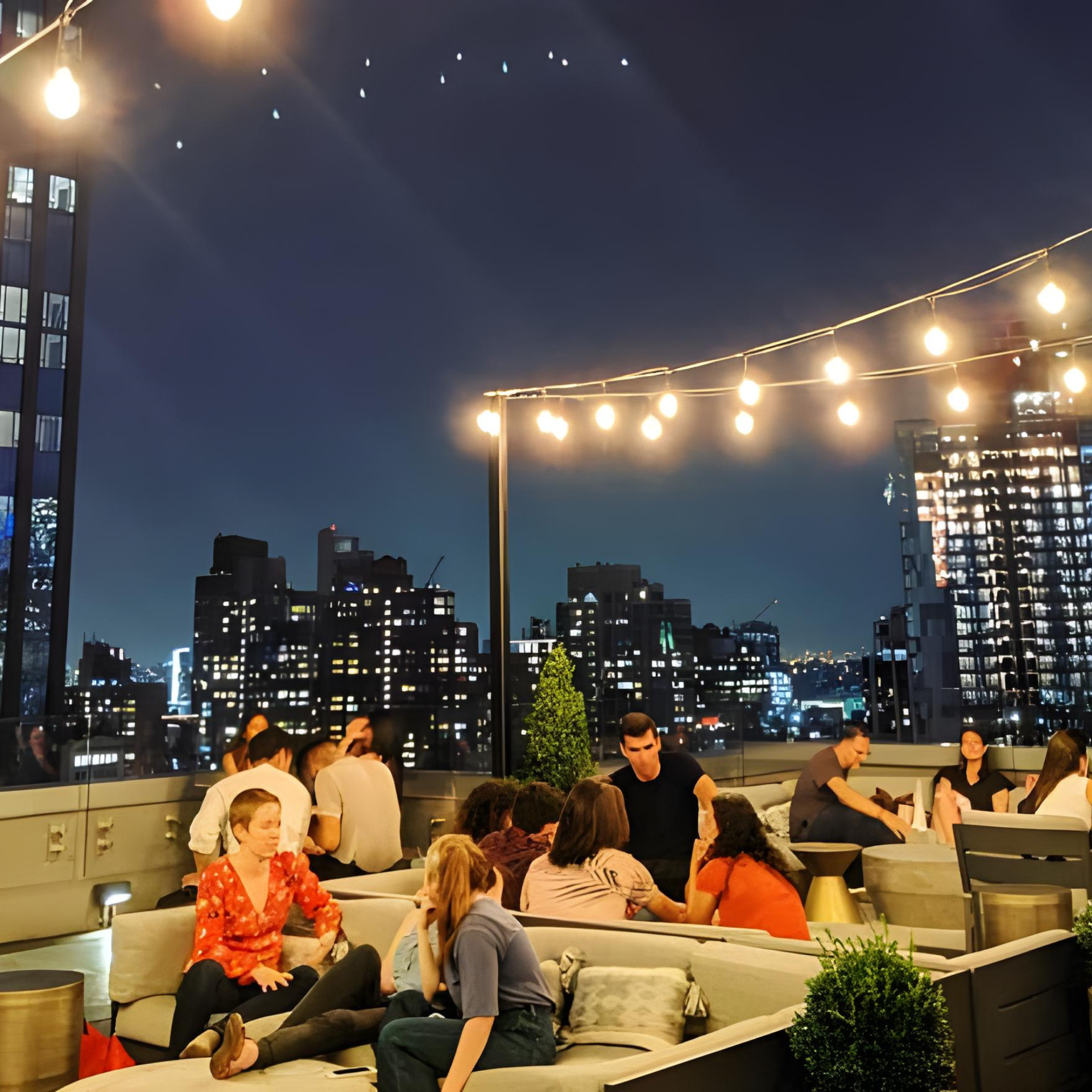 Nighttime rooftop lounge with string lights, city skyline, and groups of people relaxing
