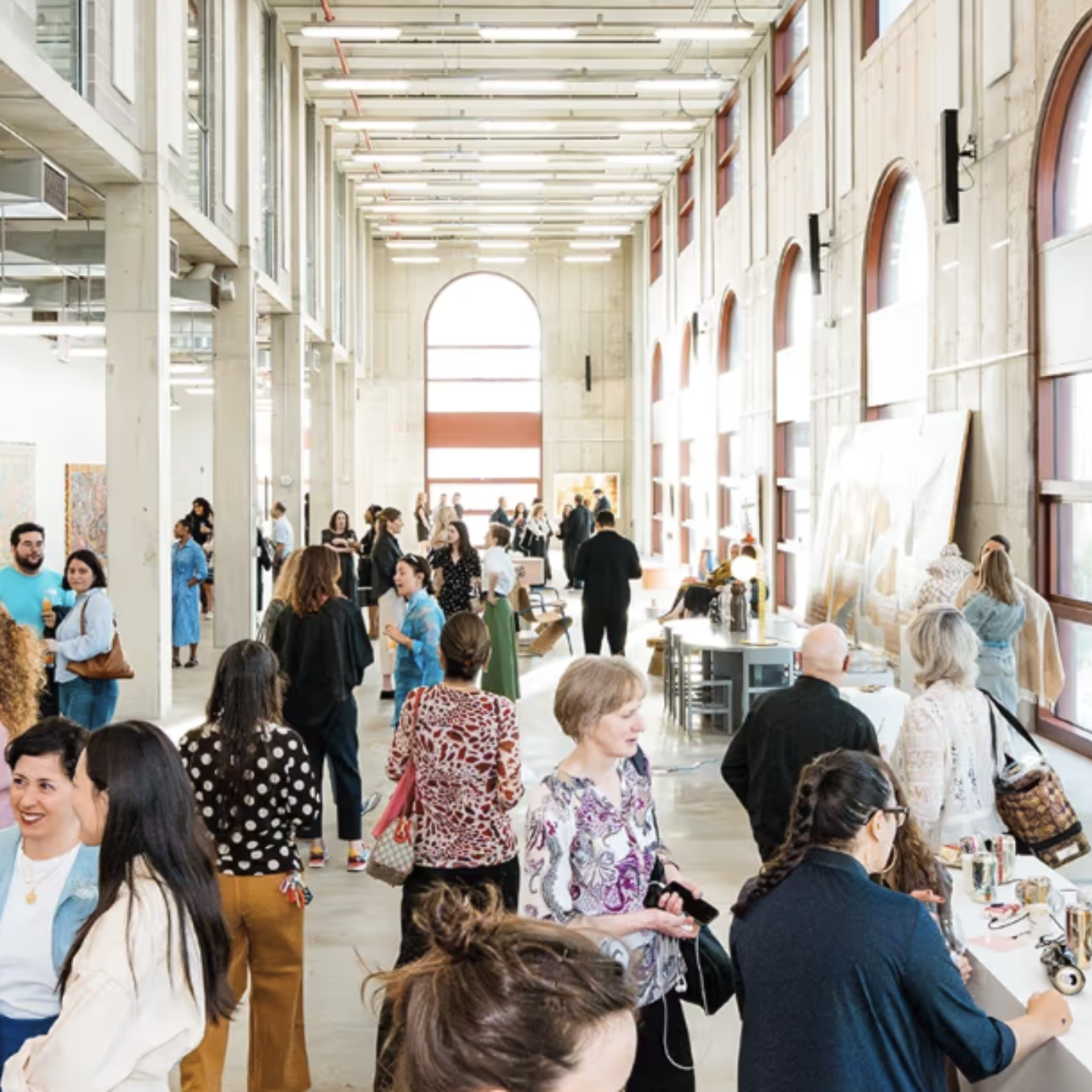 People gather in a bright gallery space with tall arched windows to view displayed artwork