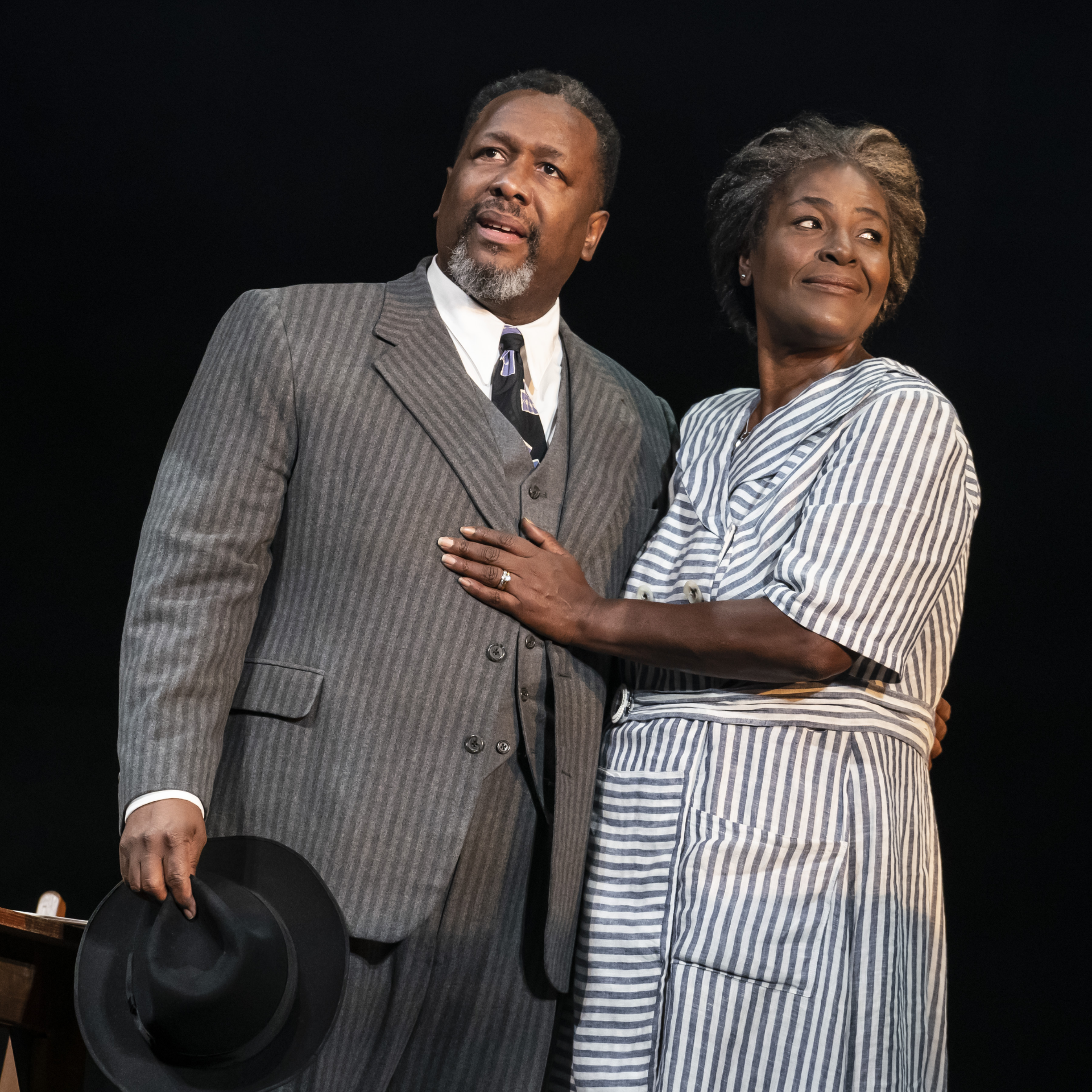 things to do in NYC: Two actors on stage in a spotlight, one in a pinstriped suit and the other in a striped dress, sharing an intimate moment