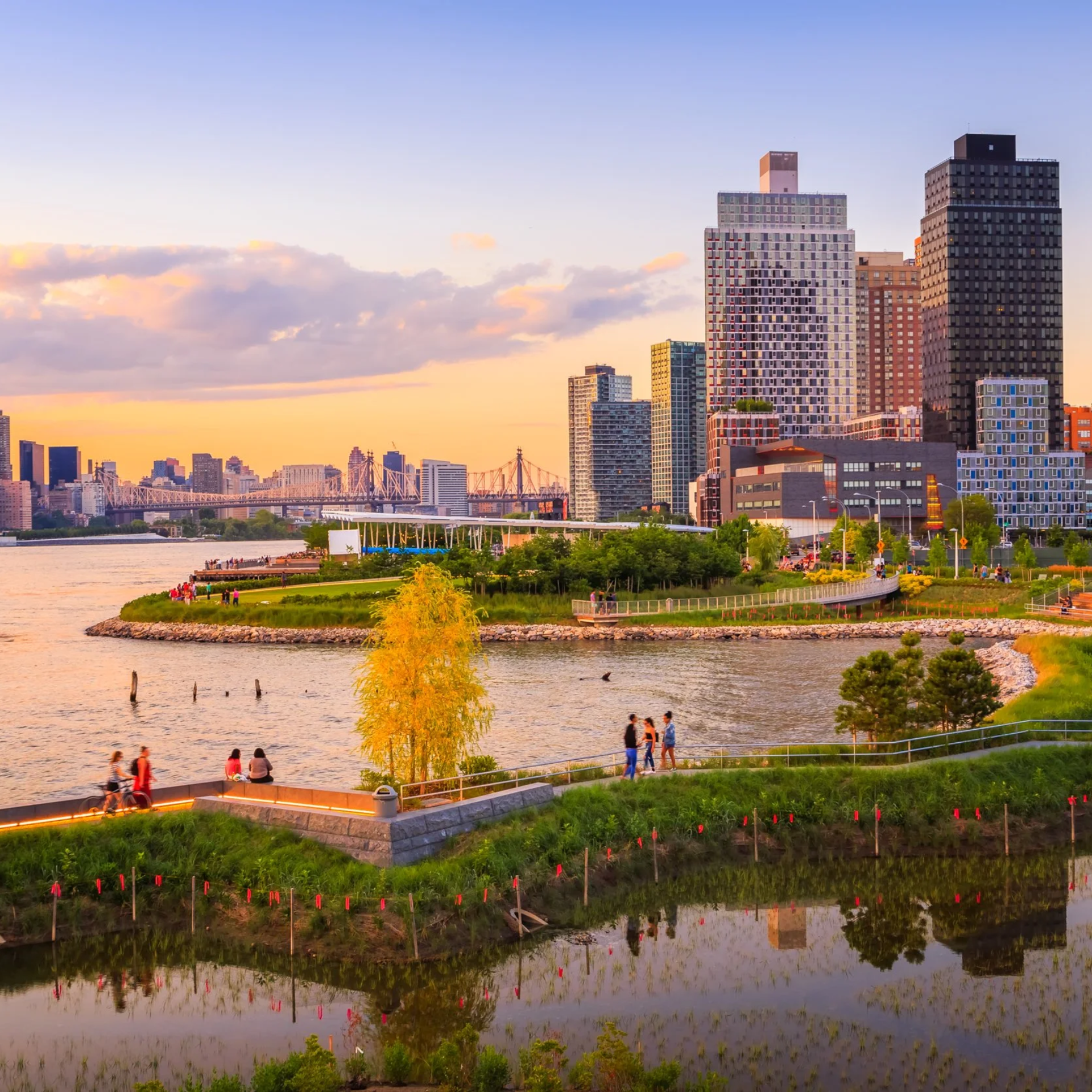 Urban waterfront park at sunset with people walking, greenery, and city skyline in the background