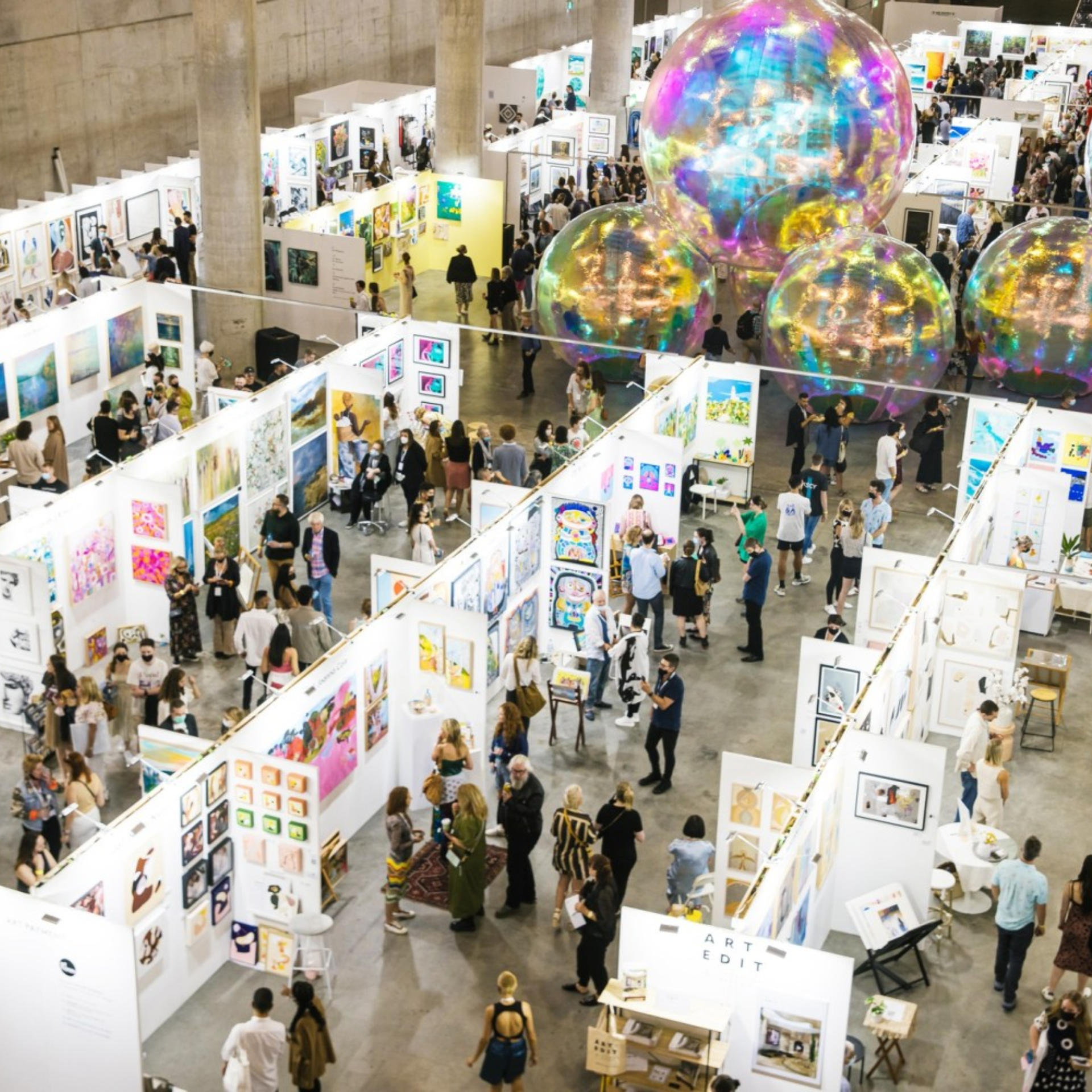 Crowds explore booths at a large indoor art fair with colorful hanging decorations above