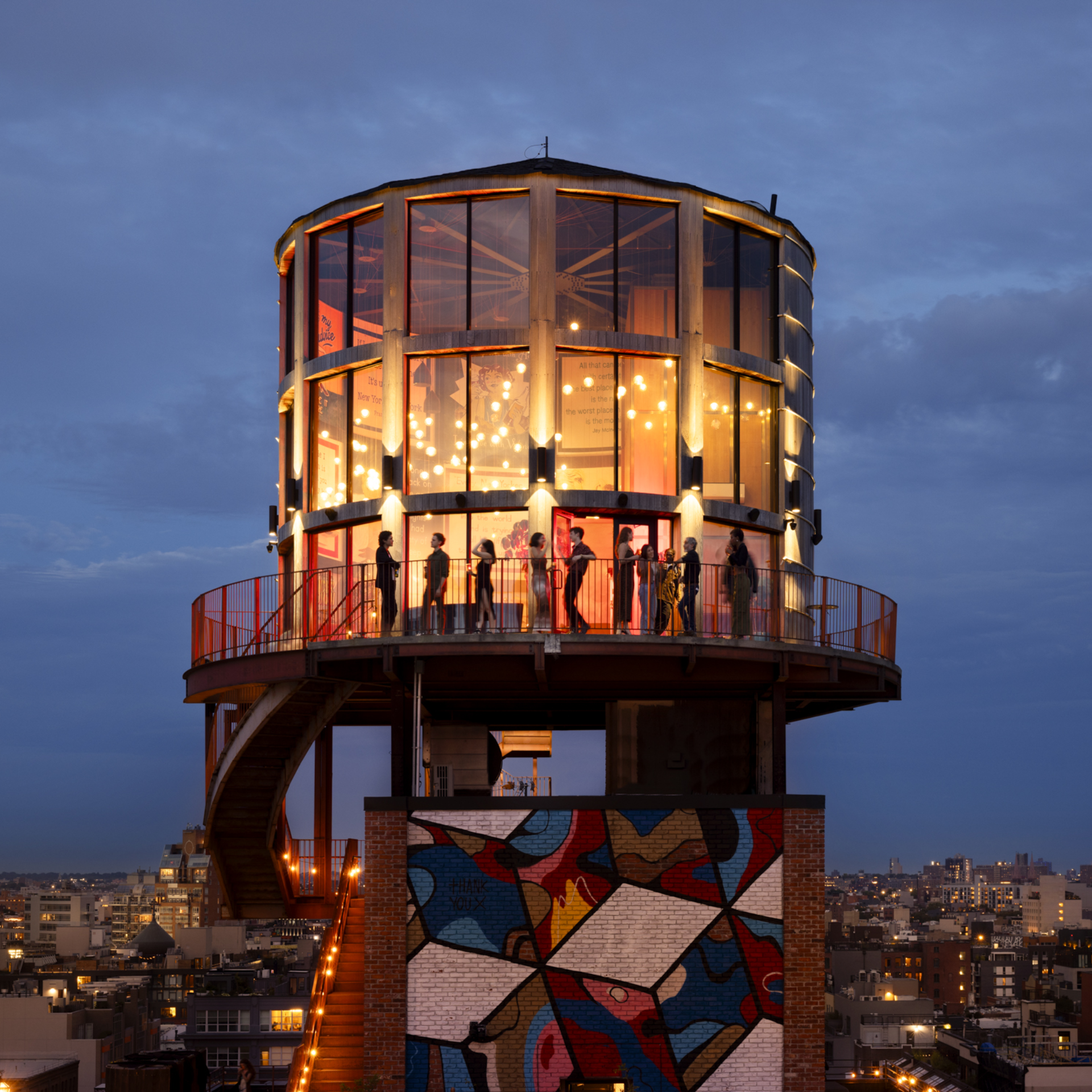 Glass cylindrical structure with balcony and mural, illuminated at dusk above city skyline