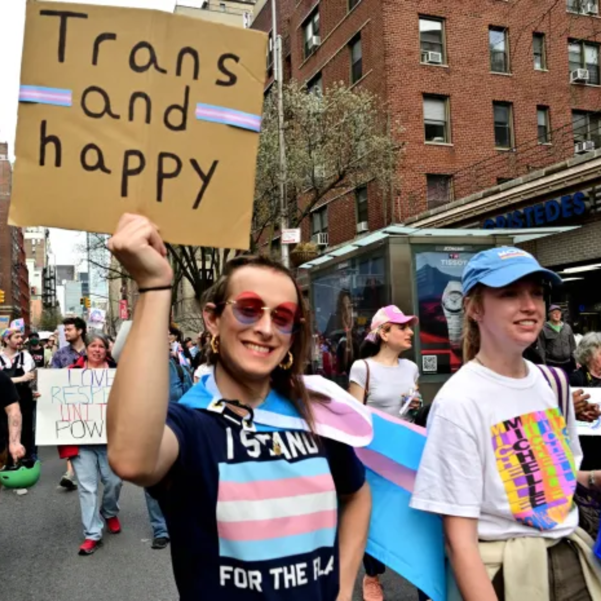People march on a city street holding signs, including one reading ‘Trans and happy