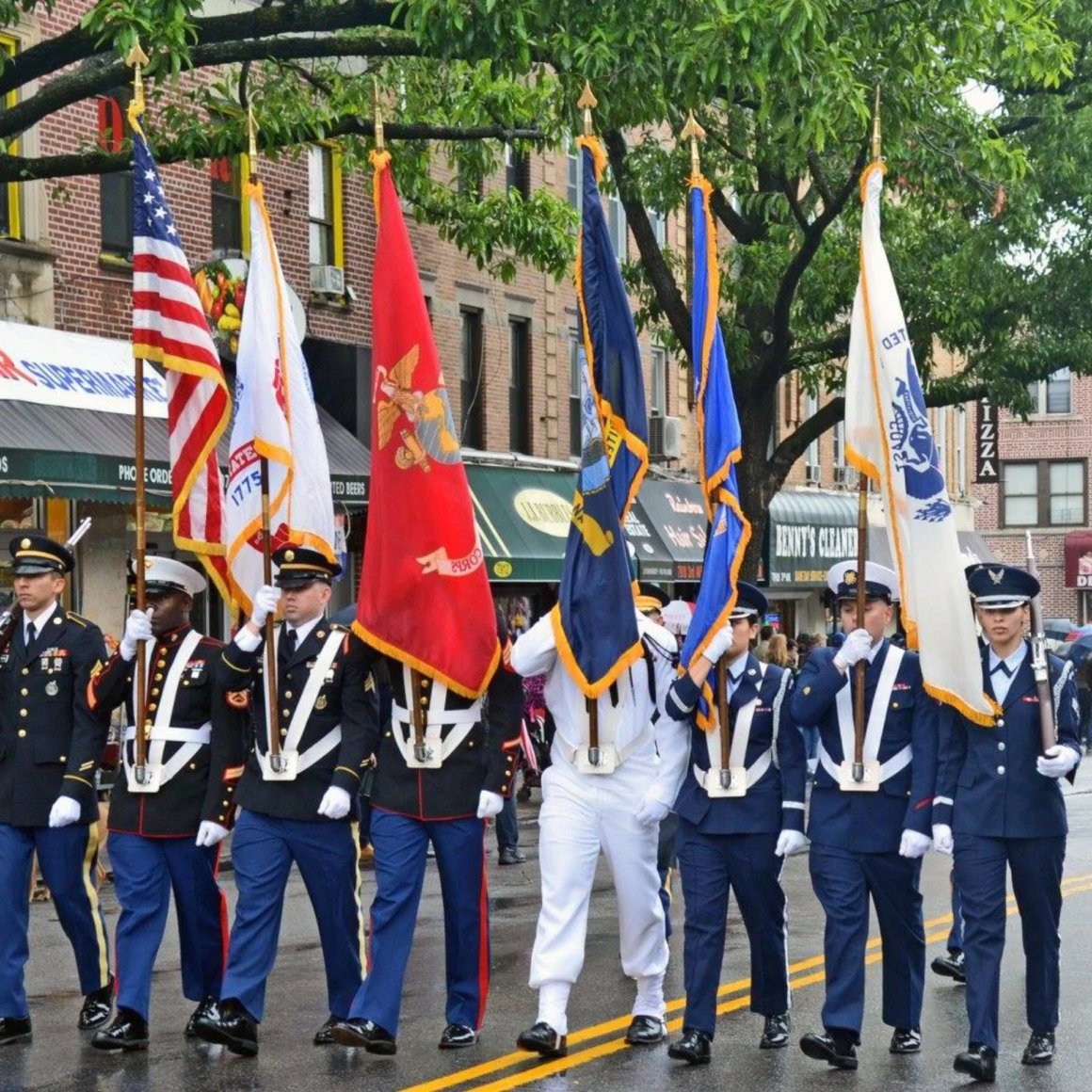 Military personnel in formal uniforms marching in city parade carrying U.S. and Armed Forces flags