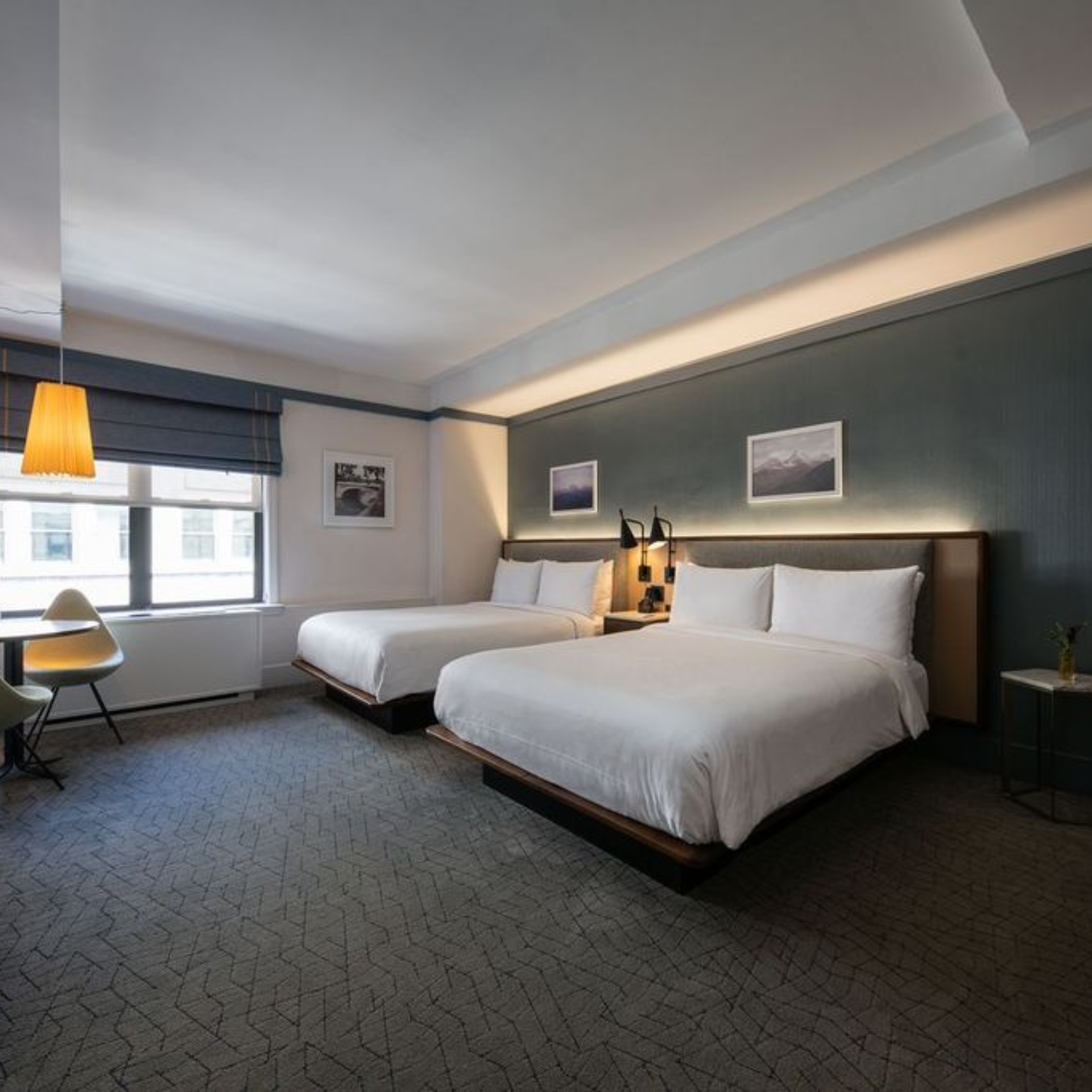 Modern hotel room with two large beds, nightstand lamps, framed photos, and seating area by window