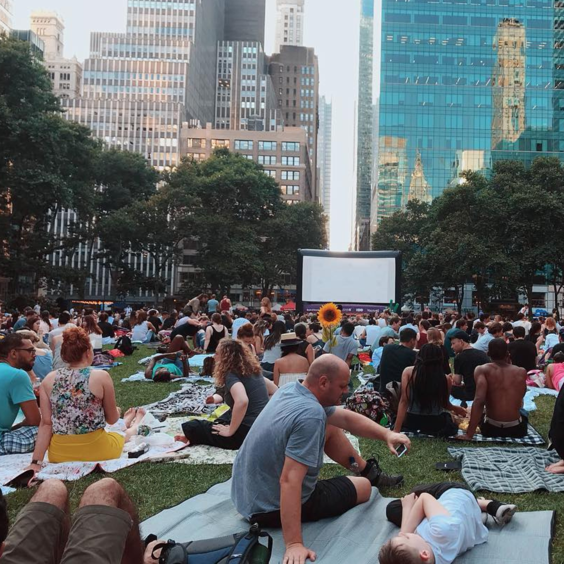A large crowd of moviegoers gathering on blankets over the green lawn for an outdoor movie night during the Bryant Park Movie Nights series in New York City, surrounded by lush trees, towering midtown Manhattan skyscrapers, and a large inflatable projection screen in the background