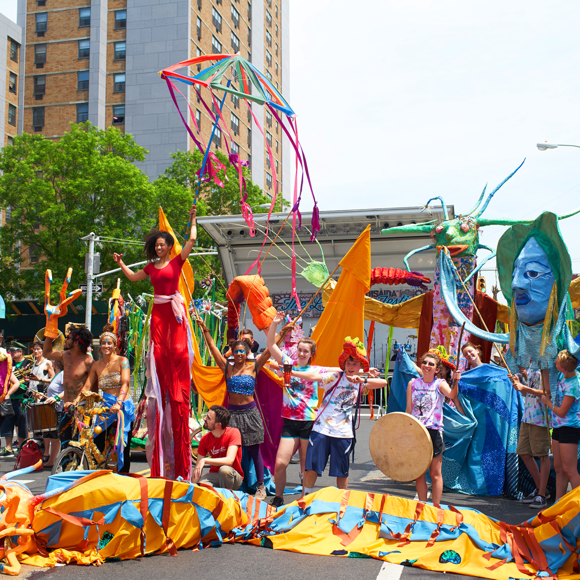 Colorful outdoor parade with stilt performer, large puppet figures, and crowd celebrating under tall buildings