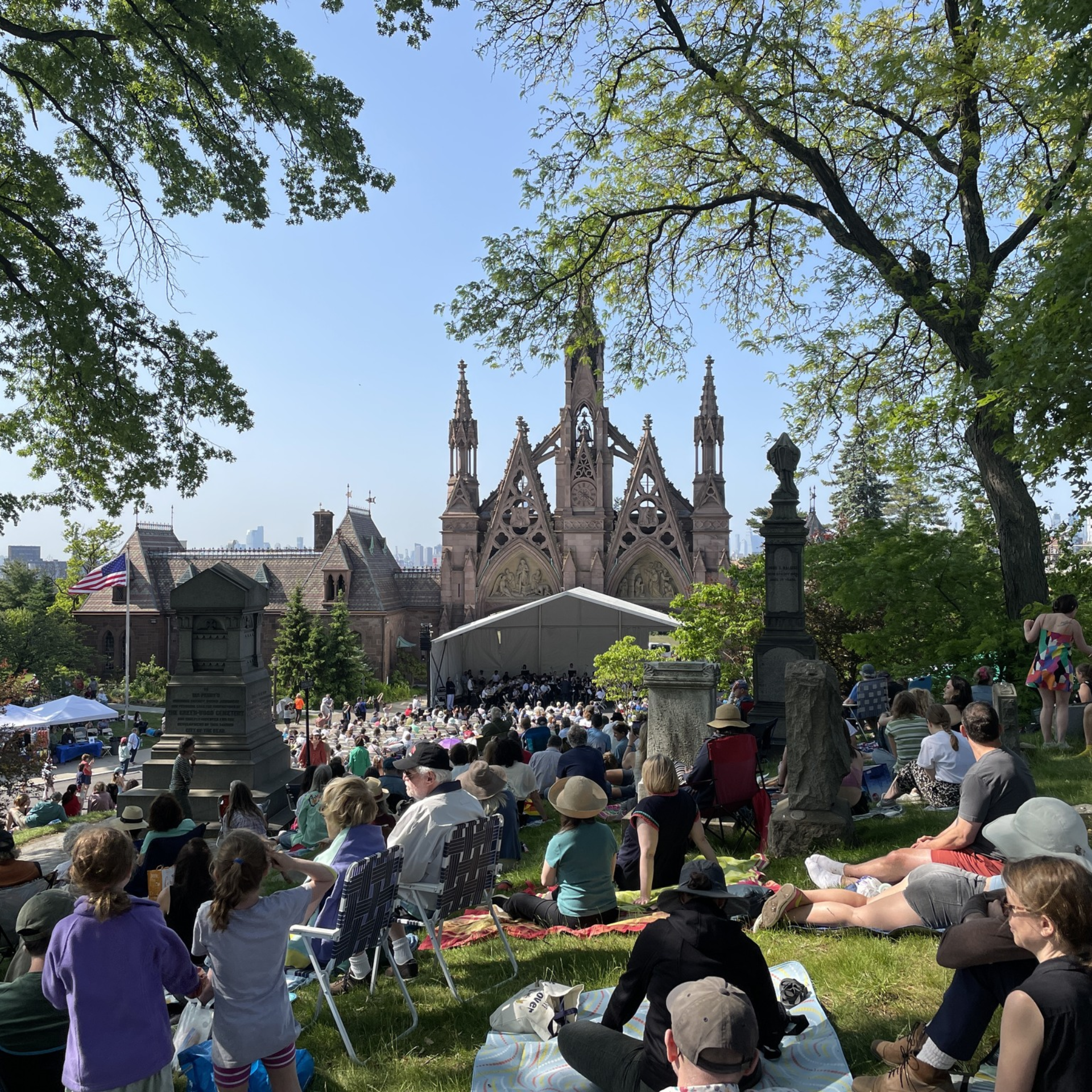 Outdoor gathering on grassy hill with crowd watching performance under white tent near ornate Gothic-style building