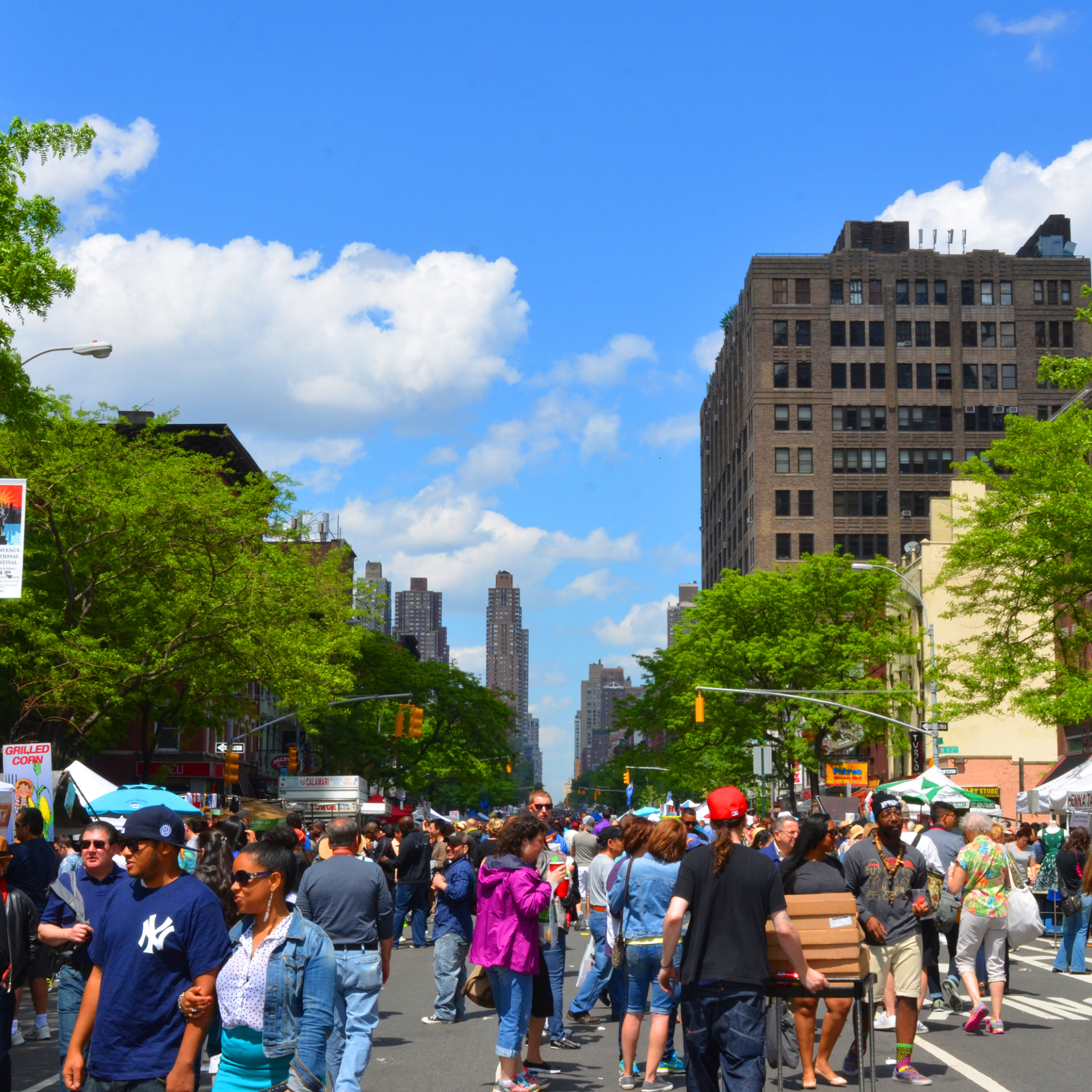 Crowded urban street fair with vendor stalls, tall buildings, and people enjoying a sunny day