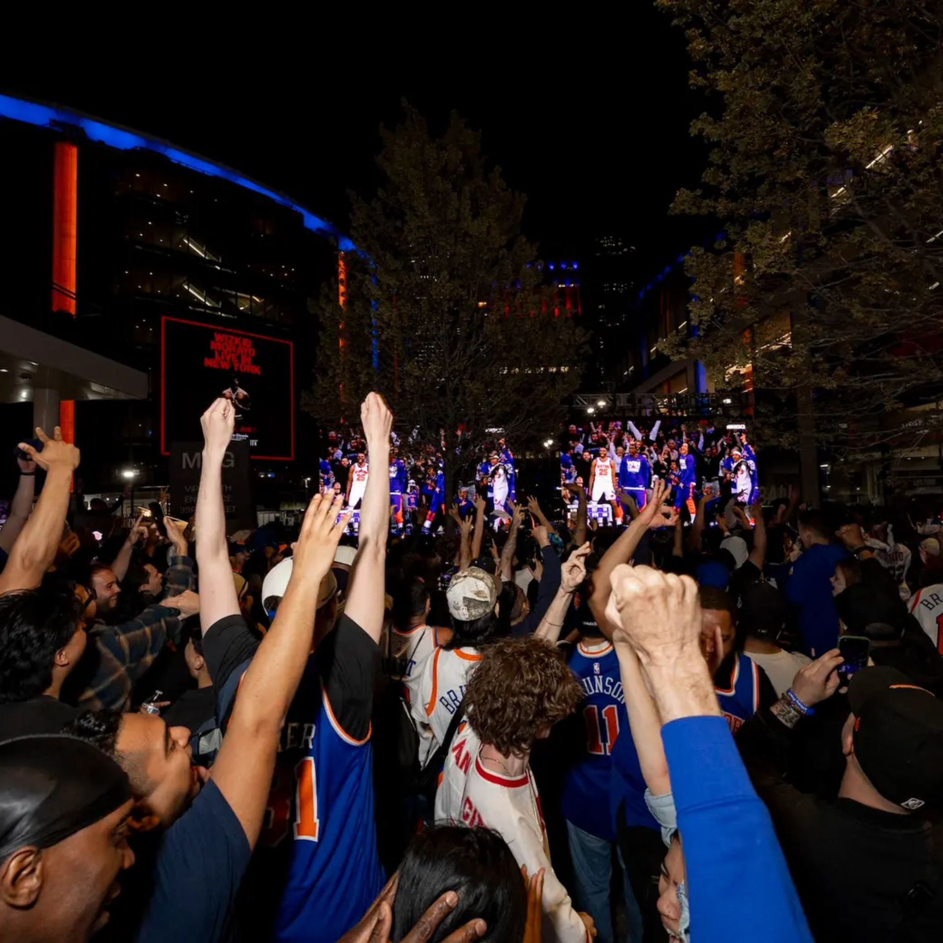 Large crowd of Knicks fans in blue and orange jerseys cheering at night with big screen and lit buildings