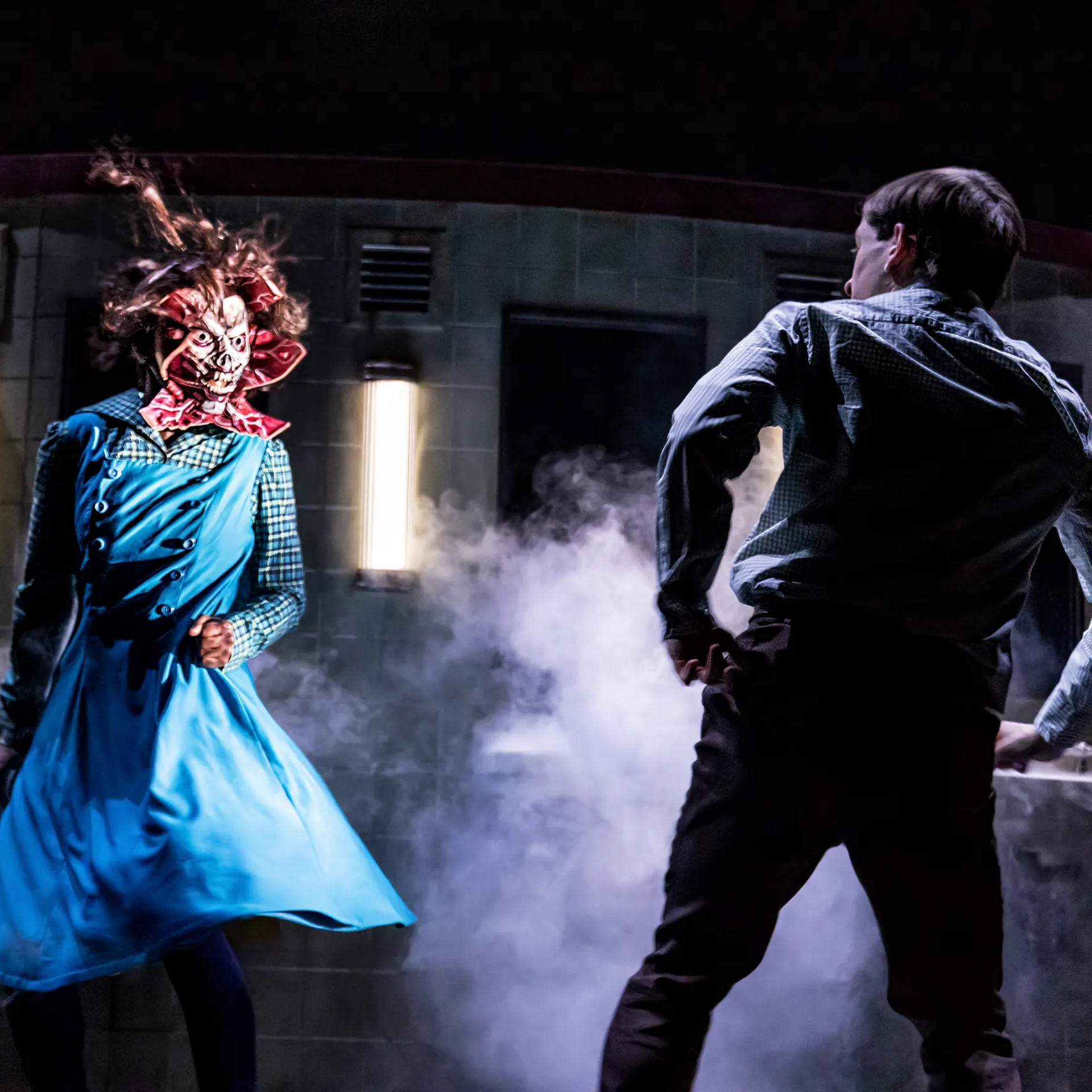 Theatrical scene with masked creature in blue dress confronting a person in checkered shirt amid smoke