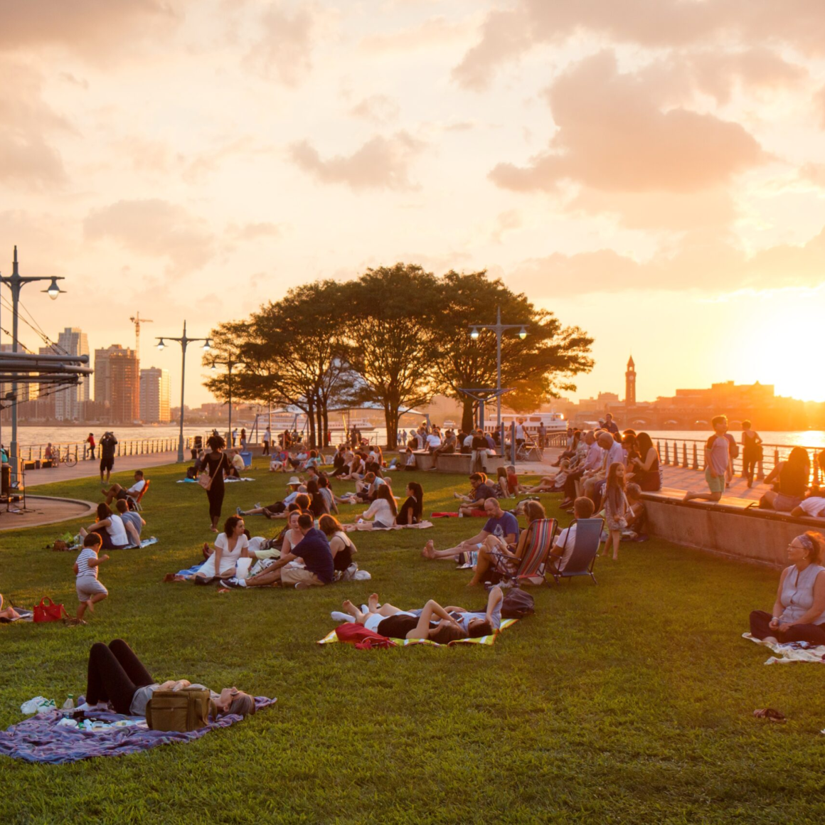 “Sunset at a waterfront park with people relaxing on the grass and city skyline in the background.