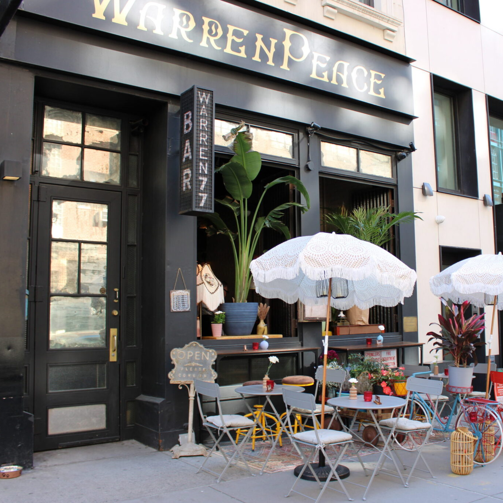 Exterior of “WarrenPeace” bar with gold-letter sign, potted plants, and outdoor seating under patterned umbrellas
