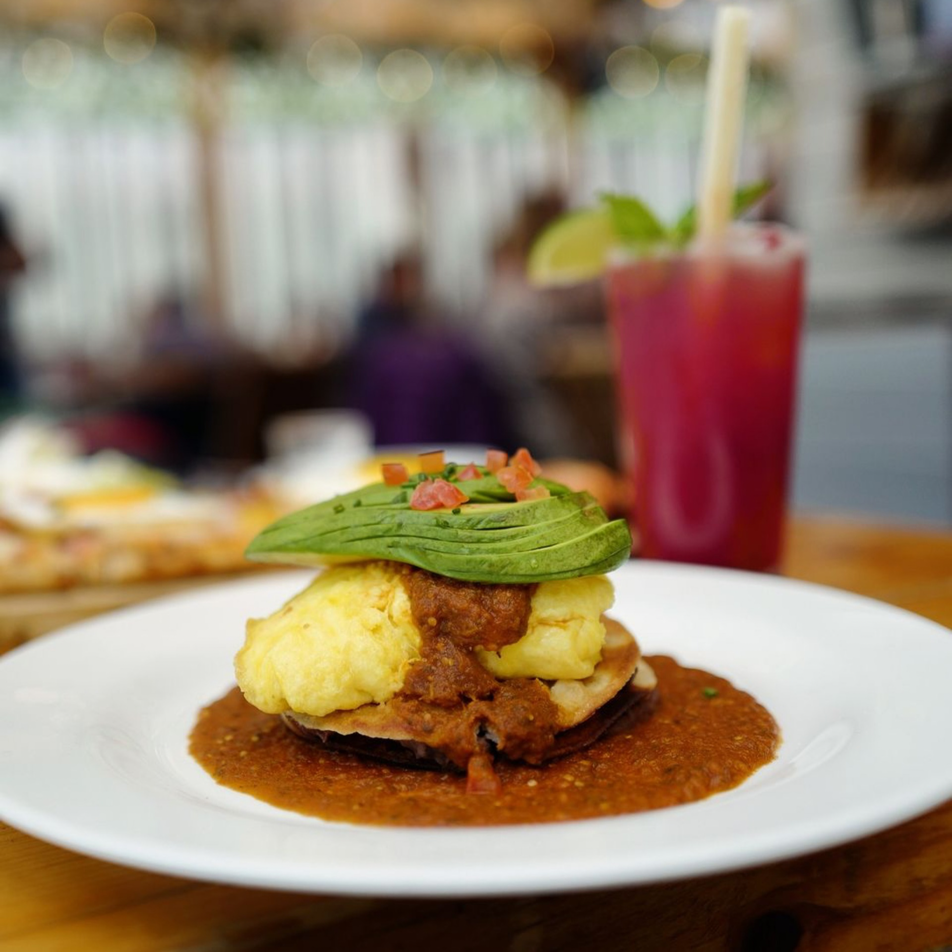 Brunch dish with tortilla base, scrambled eggs, salsa, avocado slices, and red drink on wooden table