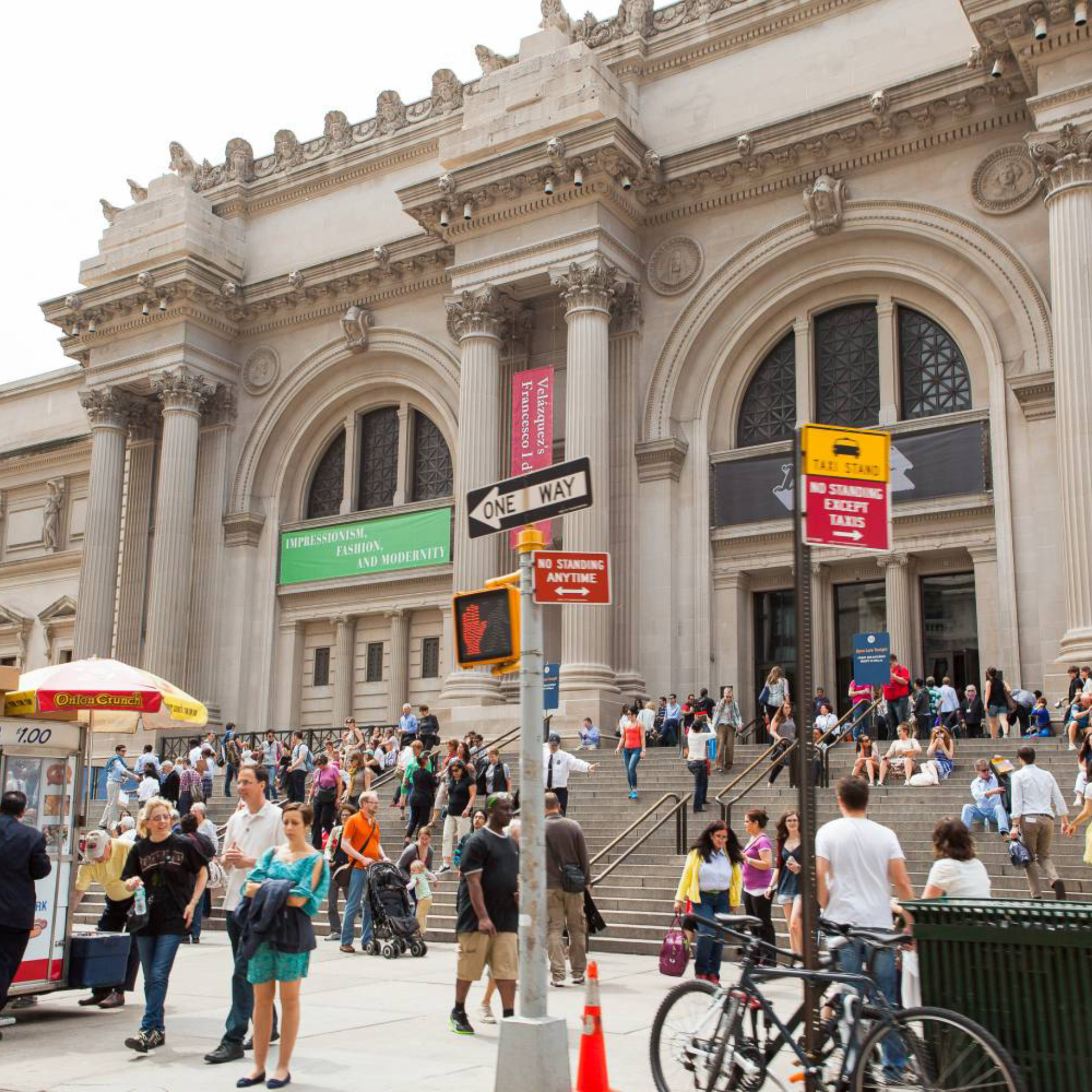 A bustling crowd of visitors gather on the iconic front steps of The Metropolitan Museum of Art (The Met) in New York City, featuring a street vendor cart, parked bicycles, and the museum's grand Beaux-Arts facade with classical columns and large arched entryways in the background