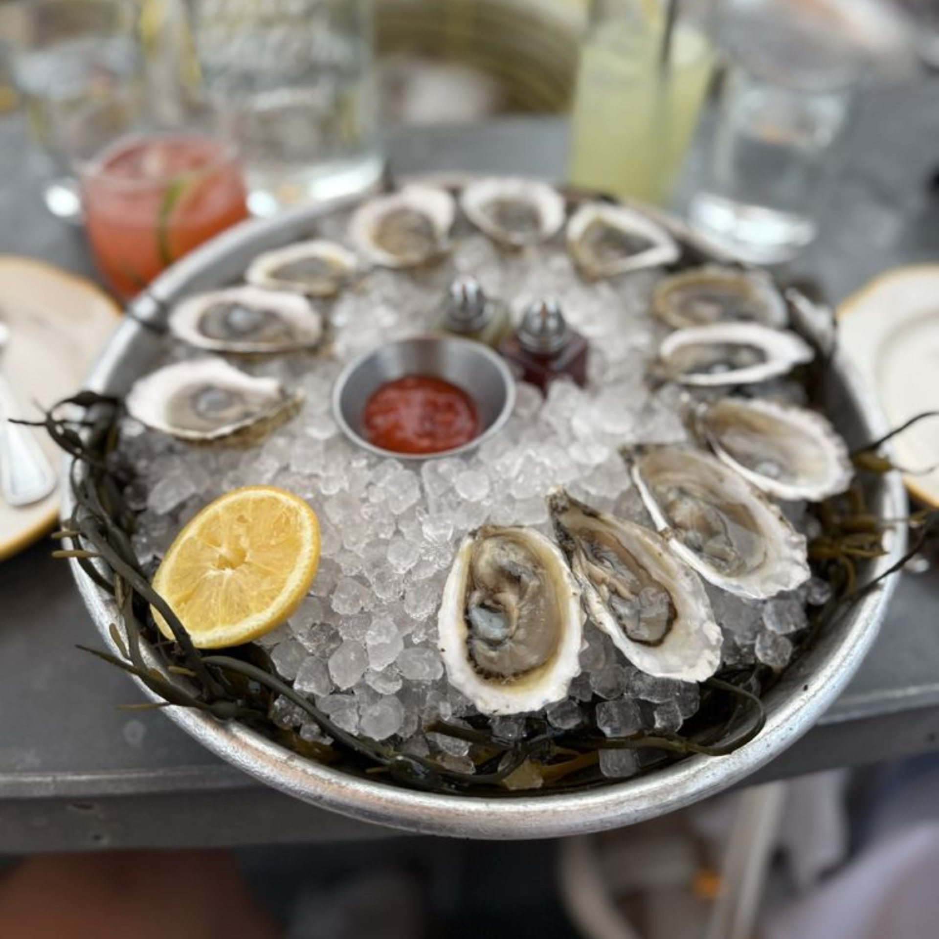 Round tray of oysters on ice with cocktail sauce, lemon, and drinks on table, NYC seafood dining experience featured in daily things to do in NYC any day