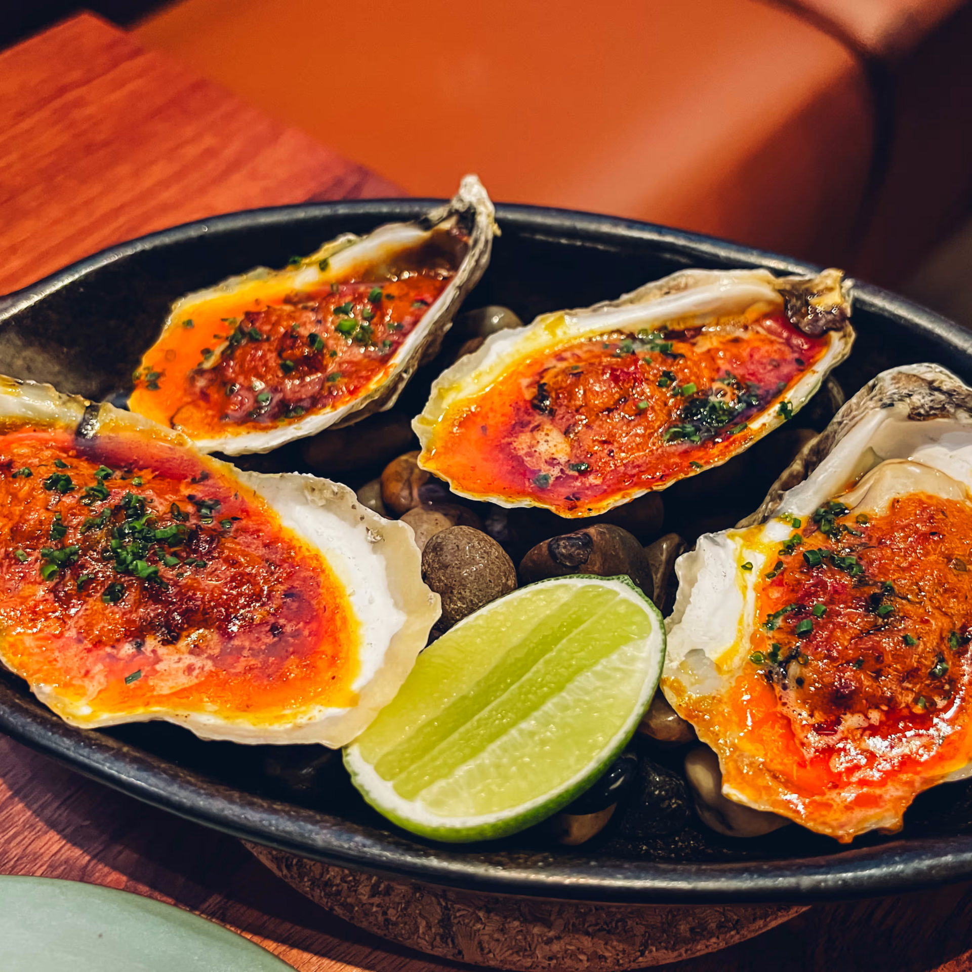 Close‑up of baked NYC oysters with reddish sauce, herb garnish, lime half, served on stones in dark dish, gourmet seafood dining featured in daily things to do in NYC any day