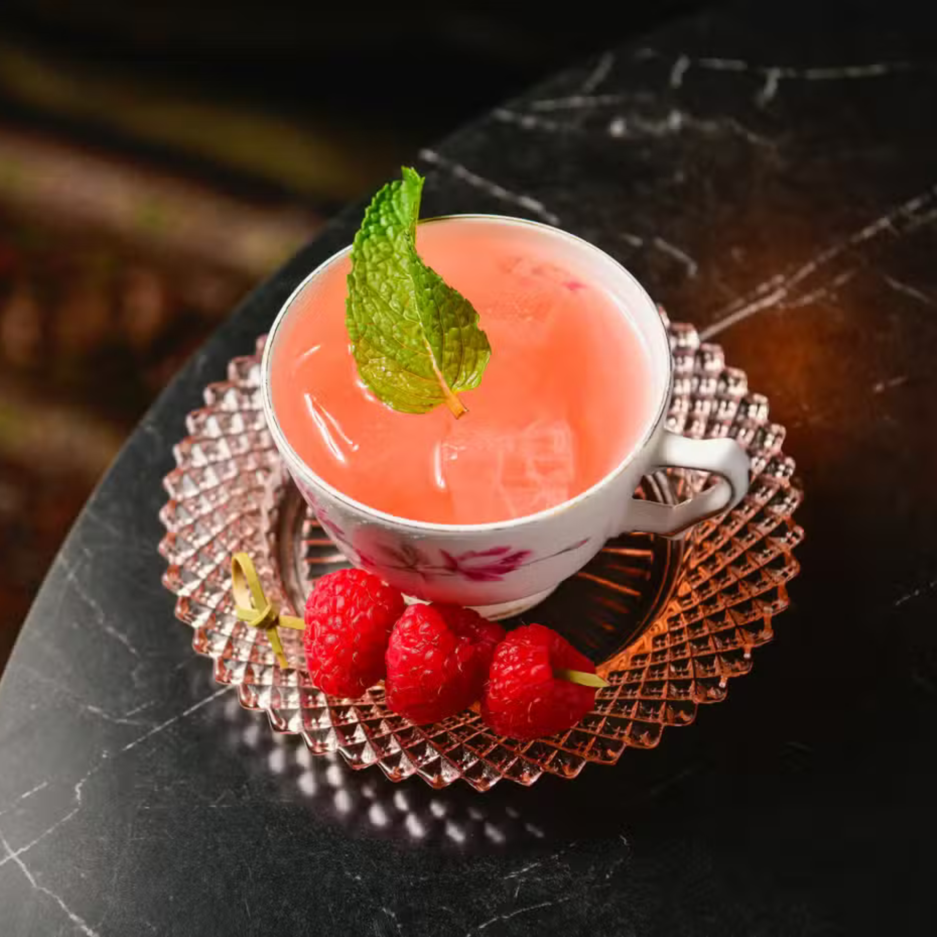 Pink drink in a teacup with mint and raspberries on a pink saucer over dark marble