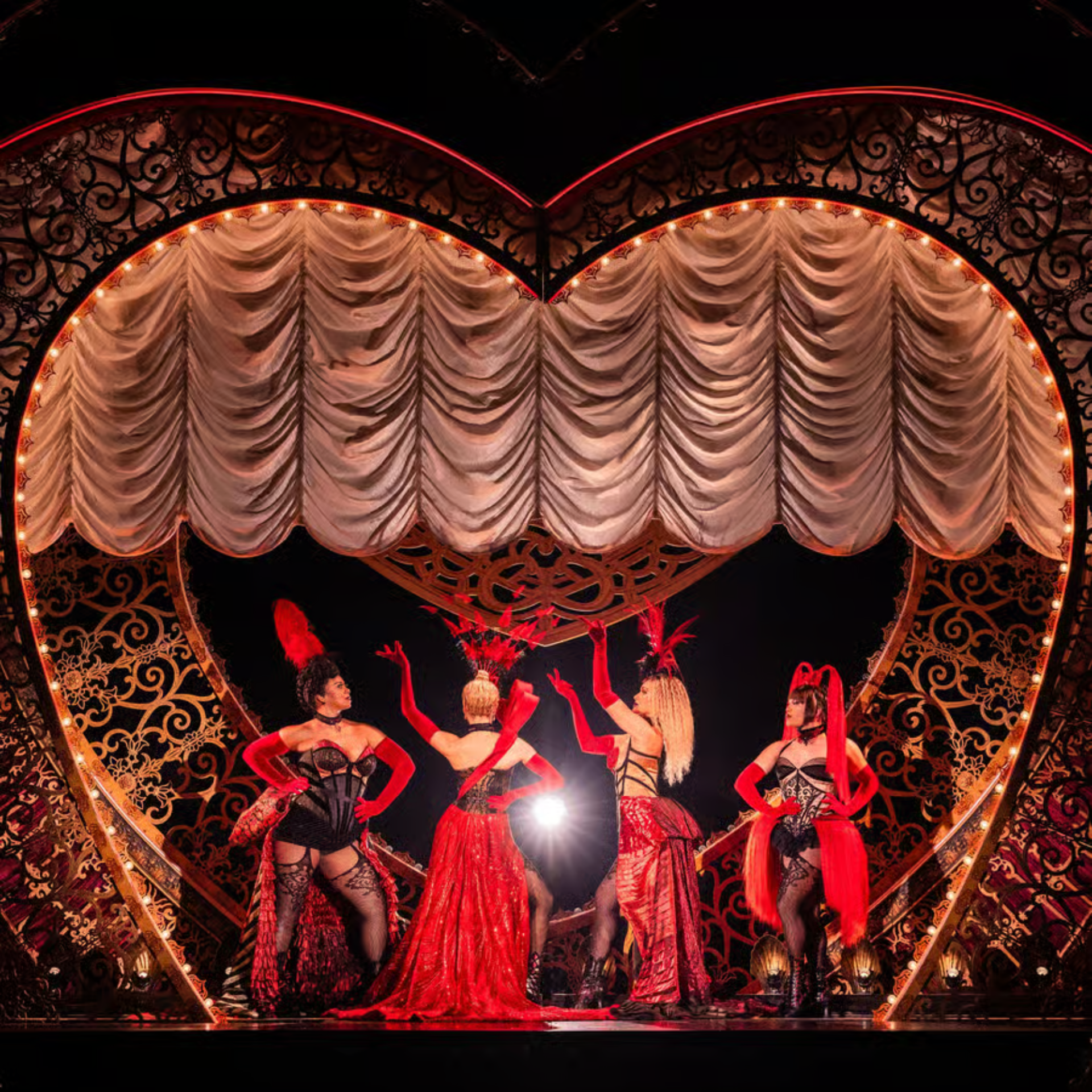 Four burlesque performers pose dramatically onstage in red and black costumes, framed by a glowing heart