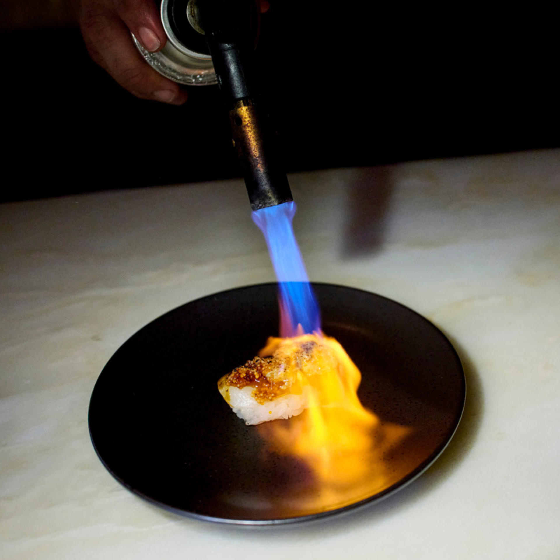 A hand sears a piece of sushi with a blue flame from a culinary blowtorch