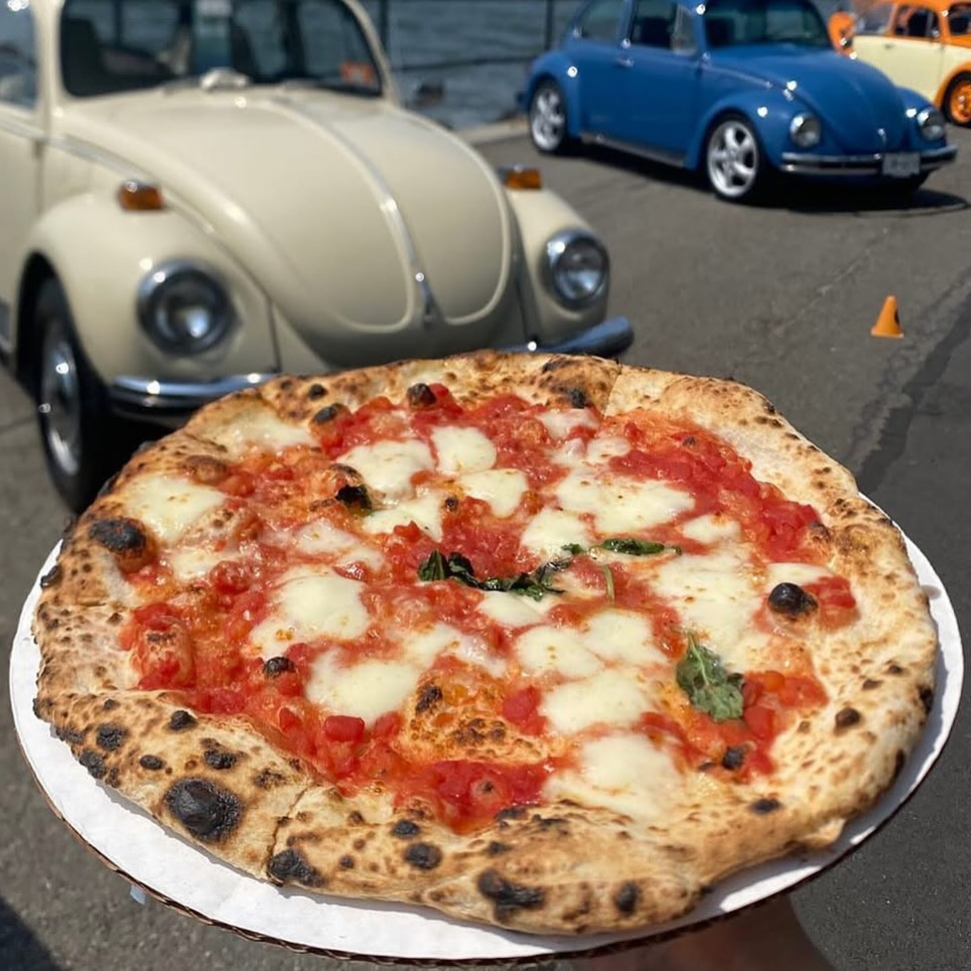 Fresh Neapolitan-style pizza held in front of three vintage Volkswagen Beetle cars