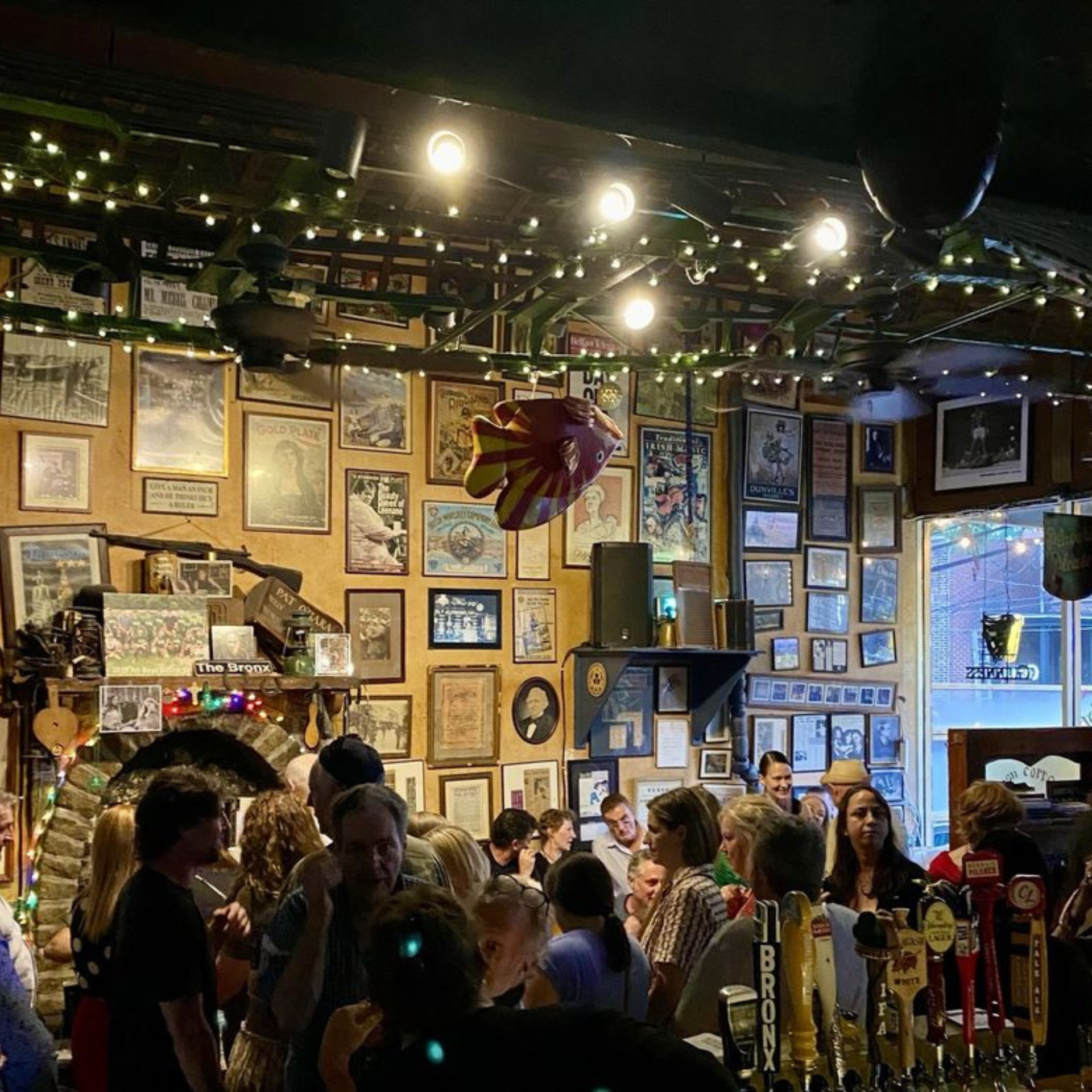 Crowded pub interior with eclectic wall décor, string lights, and beer taps at the bar