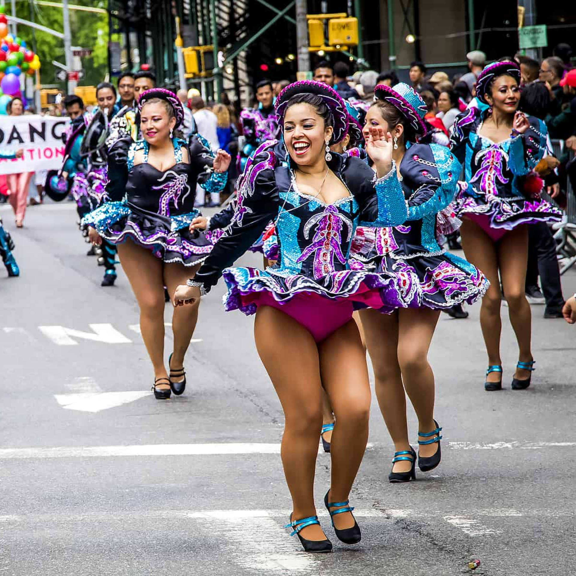 A group of smiling women in vibrant, purple and blue sequined Caporales-style costumes dance in a street parade. They wear short, ruffled skirts and matching hats, while a crowd and city buildings are visible in the background