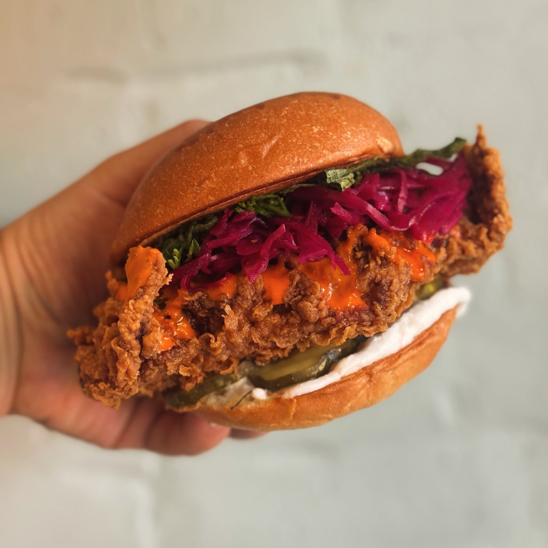 Hand holding a fried chicken sandwich with pickled cabbage, herbs, sauces, and a brioche bun