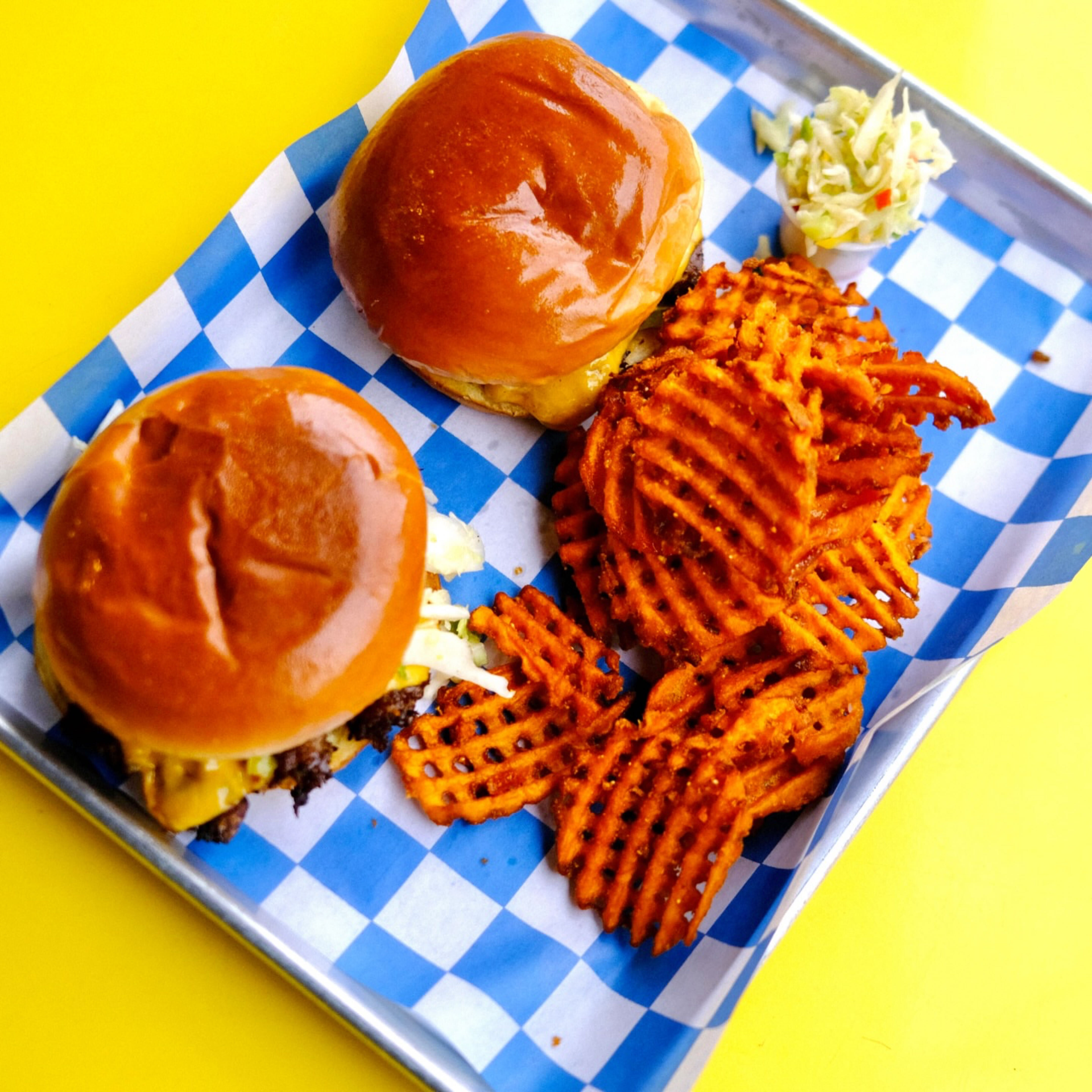 Tray with two cheeseburgers, sweet potato waffle fries, and coleslaw on blue checkered paper