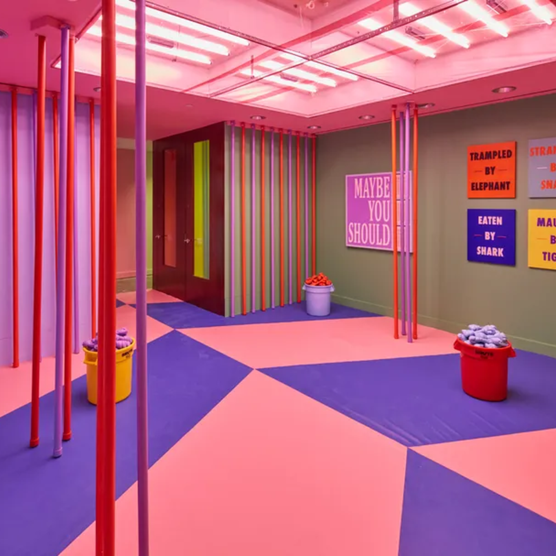 A vibrant, pink and purple art installation room featuring colorful vertical poles, geometric floor patterns, and wall signs with text like "MAYBE YOU SHOULD" and "EATEN BY SHARK