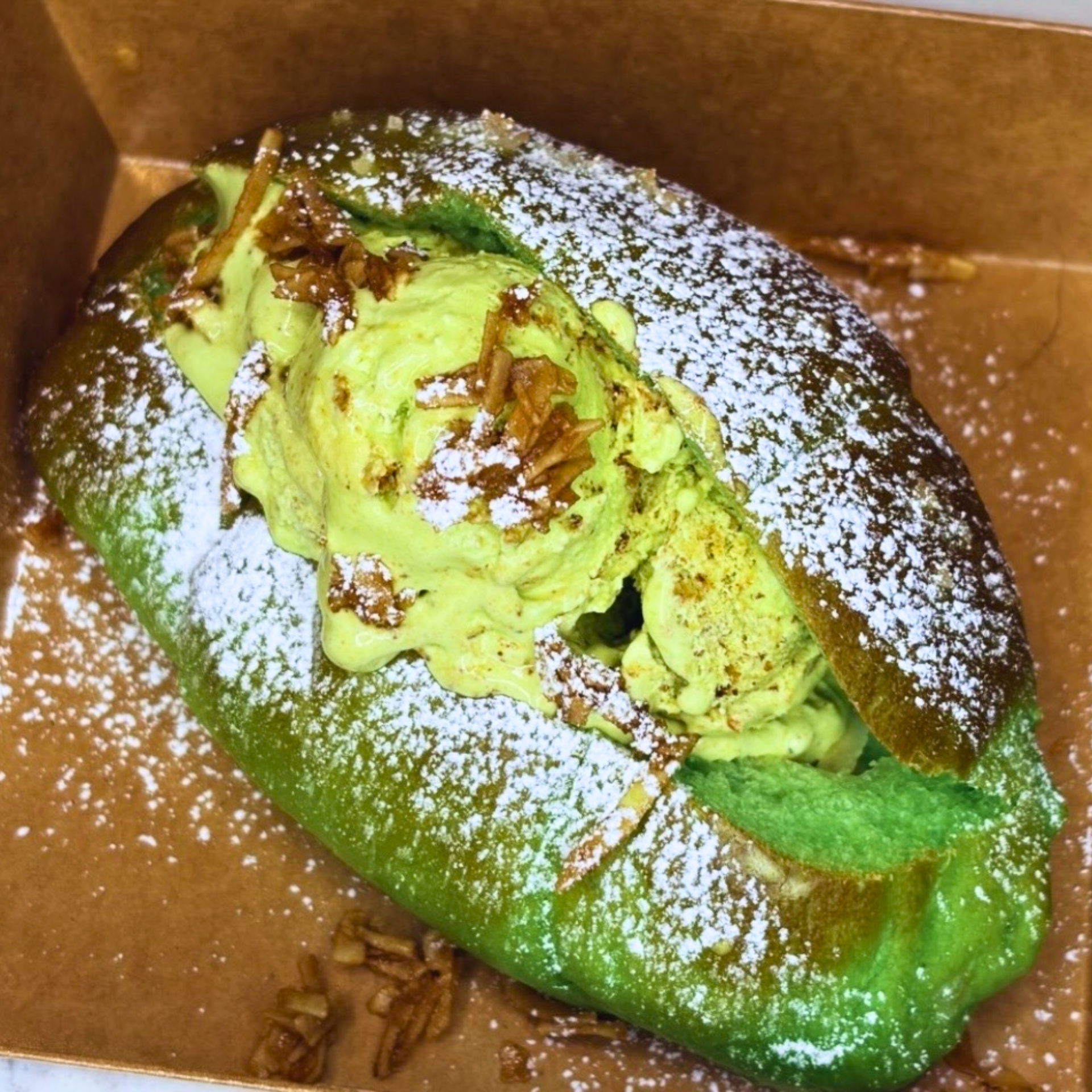 Green bread roll filled with green ice cream, topped with coconut and powdered sugar in a cardboard tray