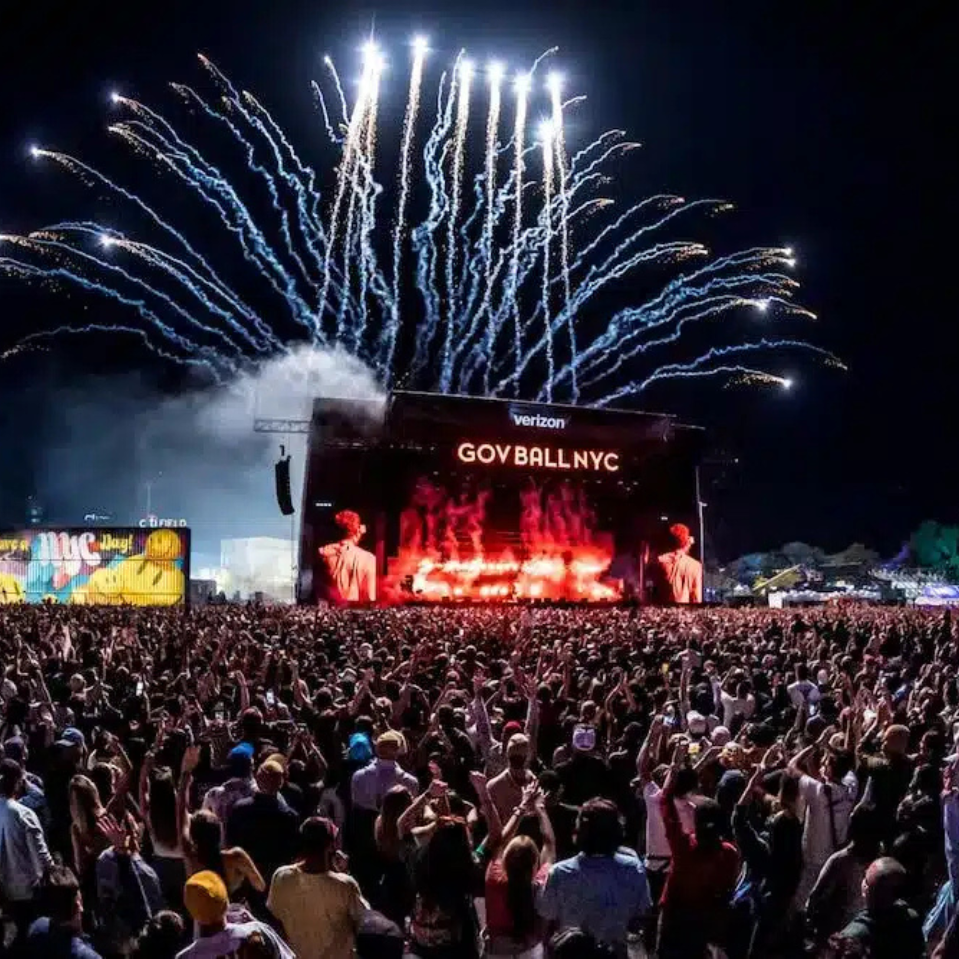 A massive crowd of festival attendees with their hands raised at the Gov Ball NYC music festival at night, facing the brightly illuminated main stage sponsored by Verizon, while a dramatic display of blue and white fireworks erupts directly above the stage