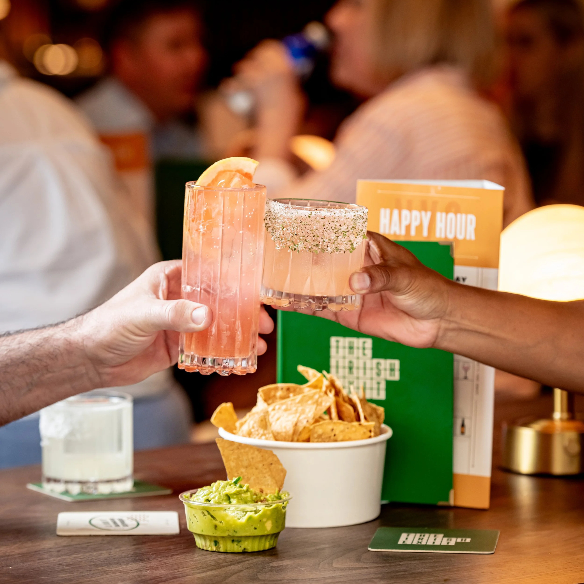 Two people clinking pink cocktails during happy hour at The Parish, with chips and guacamole on the table